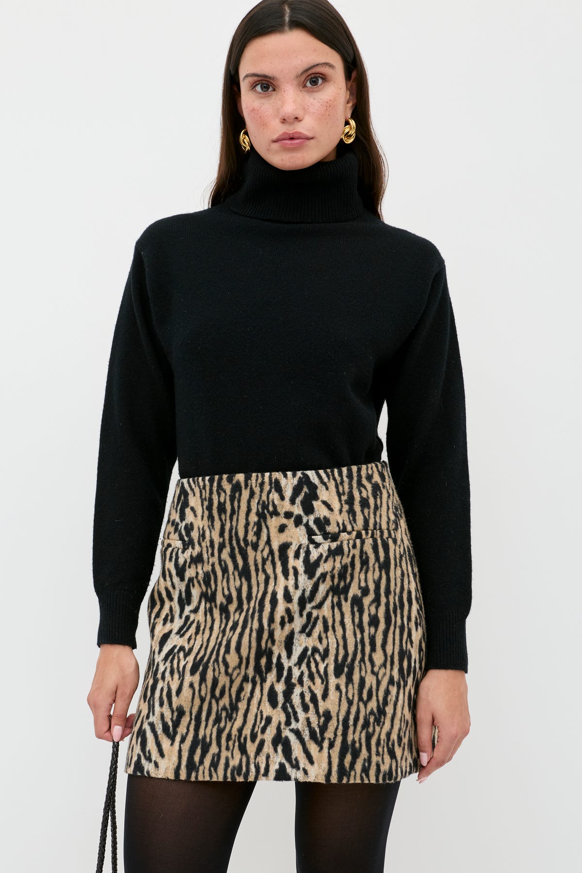 A woman in a black turtleneck, black tights, gold earrings, and the Mariah - Bohemia Leopard Brown mini skirt stands against a white background, holding a black purse—serving up chic noughties style.