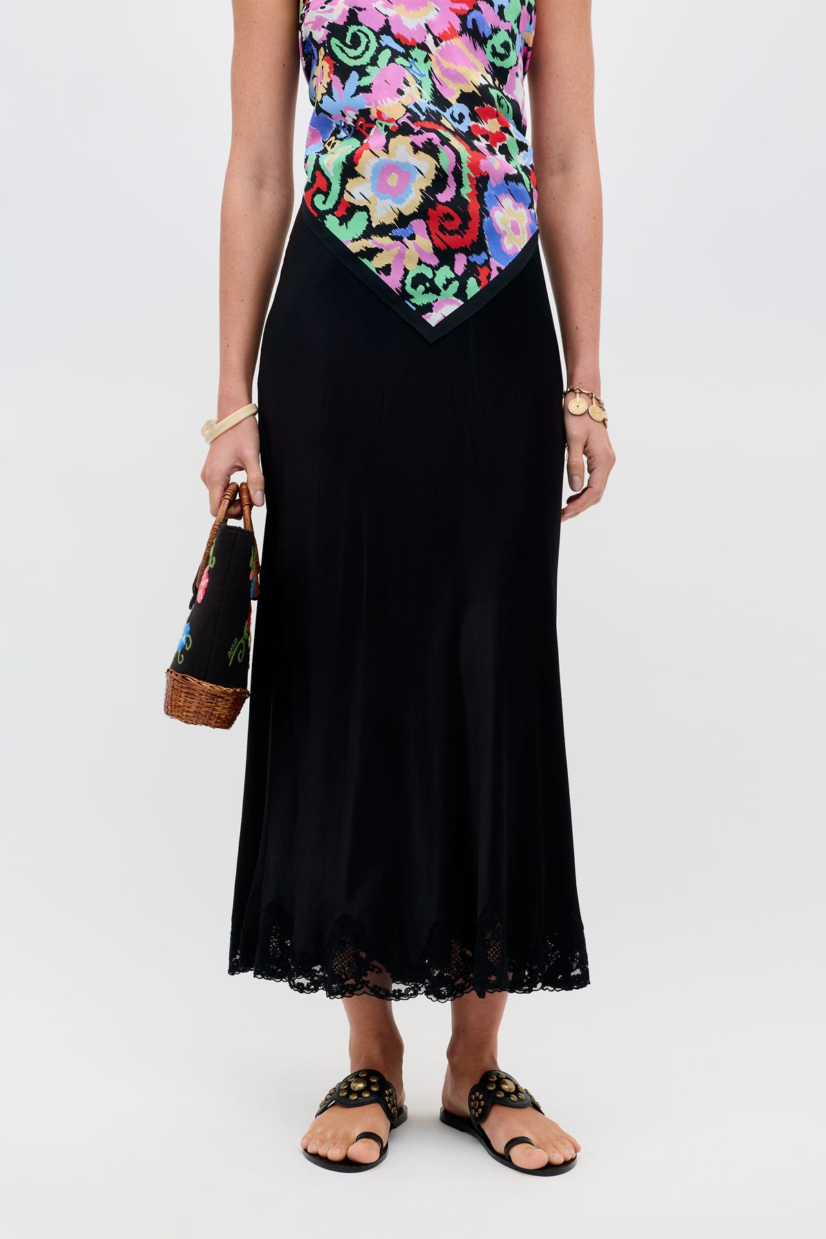 A woman wearing a colorful floral top and a black satin midi skirt with a lace hem, holding a woven handbag, and wearing the Crystal - Black sandals featuring gold details. The background is plain white.