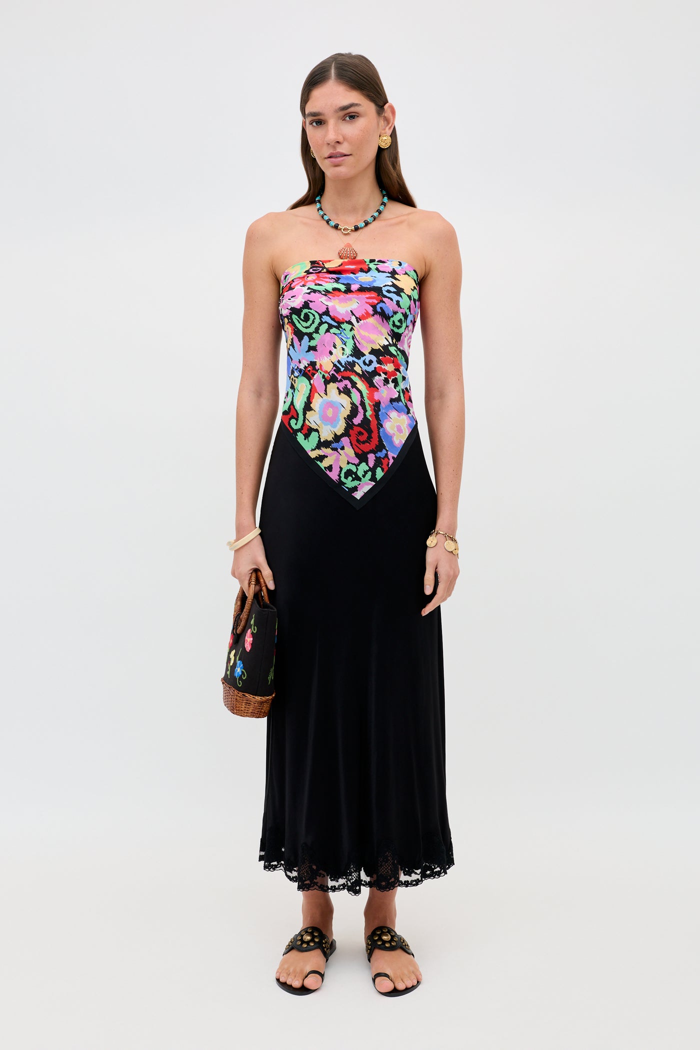 A woman wears the Crystal - Black dress, which features a colorful floral bodice and a flowing black satin midi skirt with delicate lace trim. She accessorizes with sandals, a necklace, a bracelet, and a handbag while standing against a plain white background.
