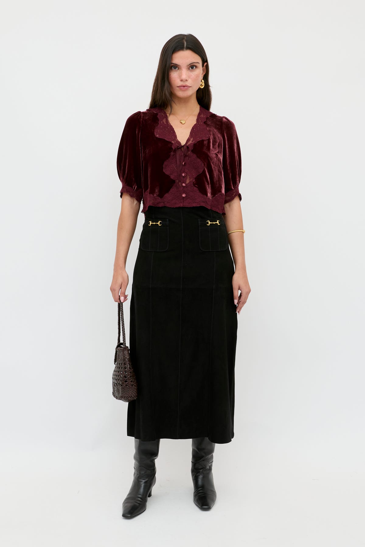 A woman with straight brown hair and a serious expression stands against a white background wearing the Edwina - Black high-waisted midi skirt with gold accents, a burgundy velvet blouse, black knee-high boots, and holding a small dark handbag.