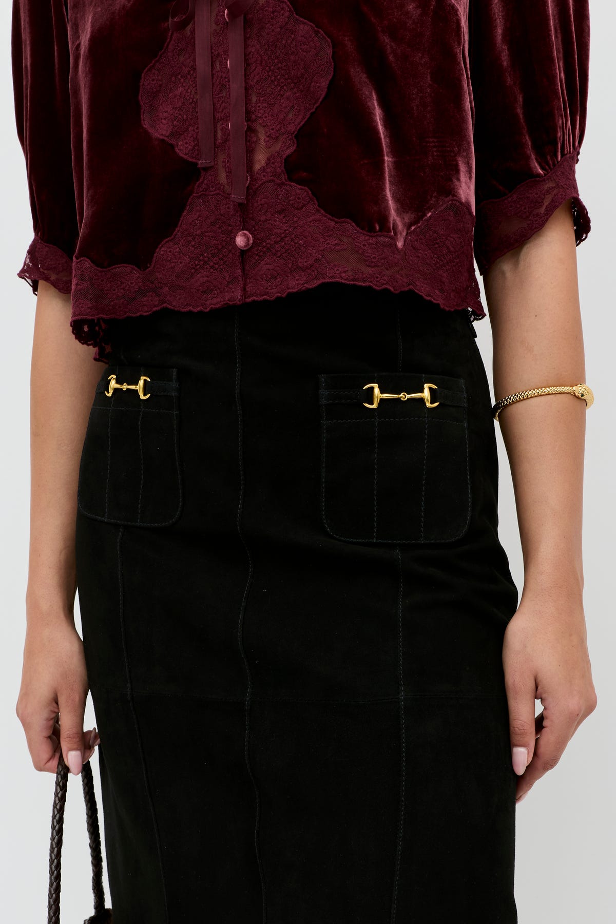A person is wearing a burgundy velvet blouse with lace trim, the Edwina - Black high-waisted skirt featuring gold decorative hardware on the pockets, and a gold bracelet. Only their torso and arms are visible.