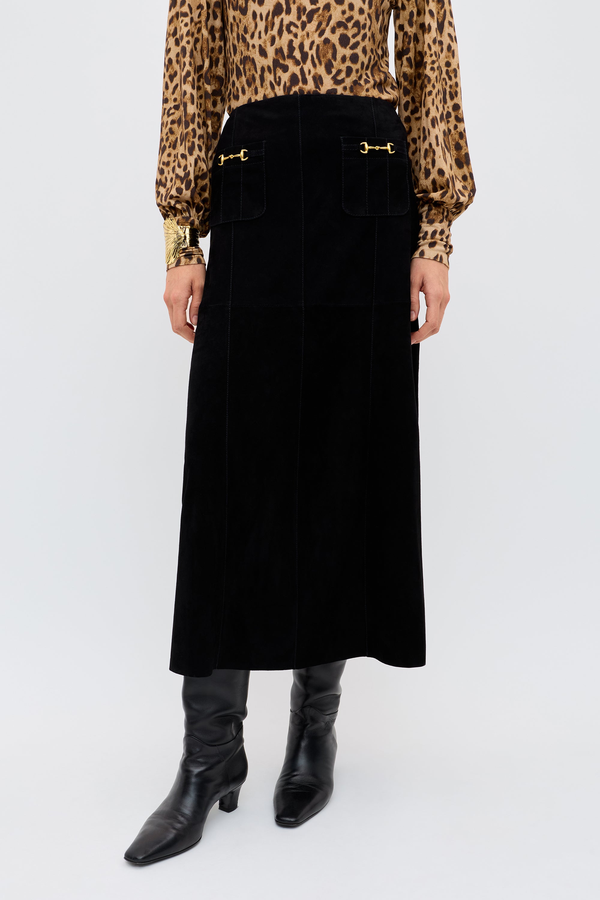 A person wearing the Edwina - Black vintage-inspired high-waisted A-line skirt with gold hardware, paired with a leopard print long-sleeve blouse and black knee-high boots, stands against a plain background.
