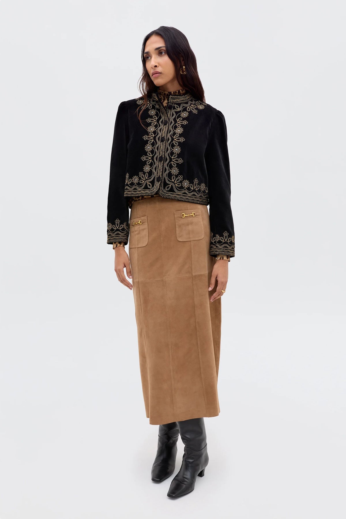 A woman stands against a plain background wearing the Edwina - Camel, a vintage-inspired camel suede skirt with gold accents, paired with a black embroidered jacket and black knee-high boots. She looks slightly to the side, arms relaxed by her sides.
