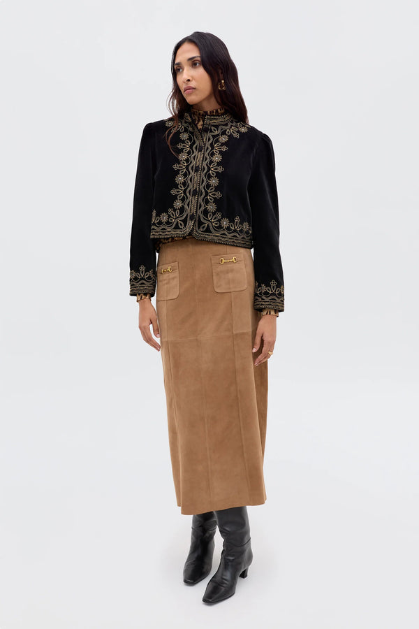 A woman stands against a plain background wearing the Edwina - Camel, a vintage-inspired camel suede skirt with gold accents, paired with a black embroidered jacket and black knee-high boots. She looks slightly to the side, arms relaxed by her sides.