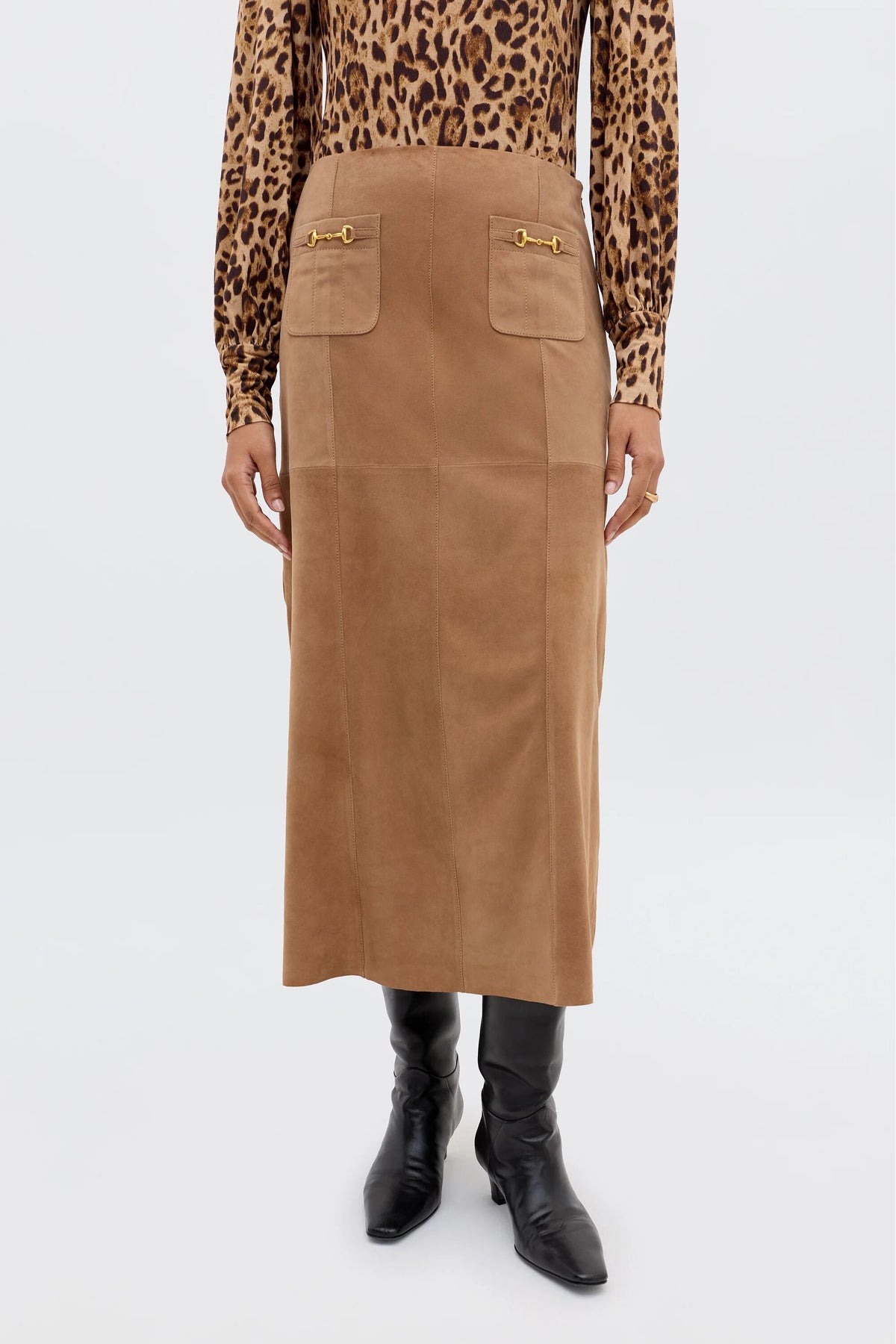 A person styles the Edwina - Camel, a vintage-inspired brown suede midi skirt with front pockets and gold accents, worn high-waisted with a leopard print blouse and black knee-high boots.
