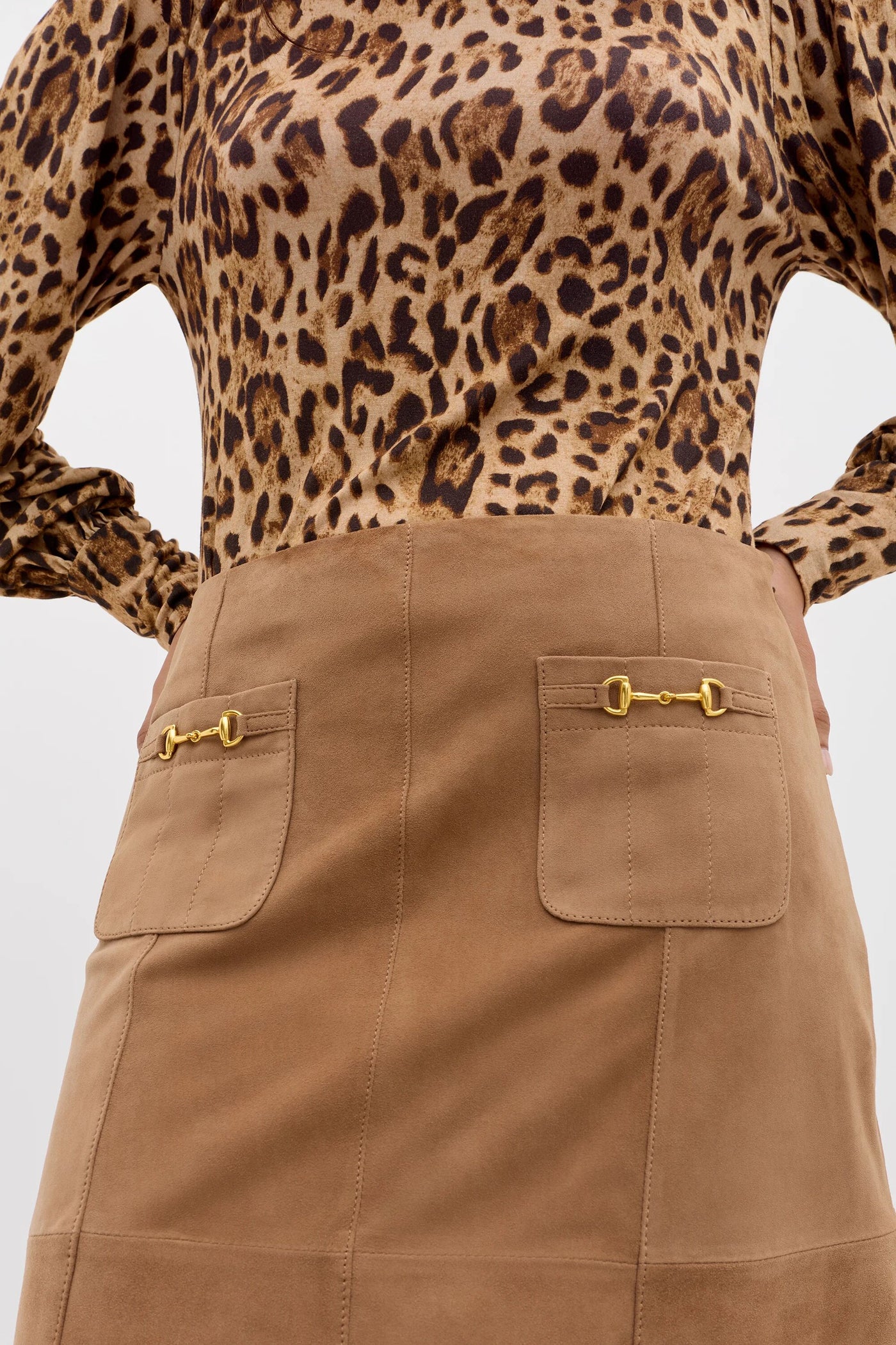 A person wears Edwina - Camel, a vintage-inspired high-waisted suede skirt with two front pockets, each adorned with a small gold buckle, paired with a leopard print long-sleeved blouse.
