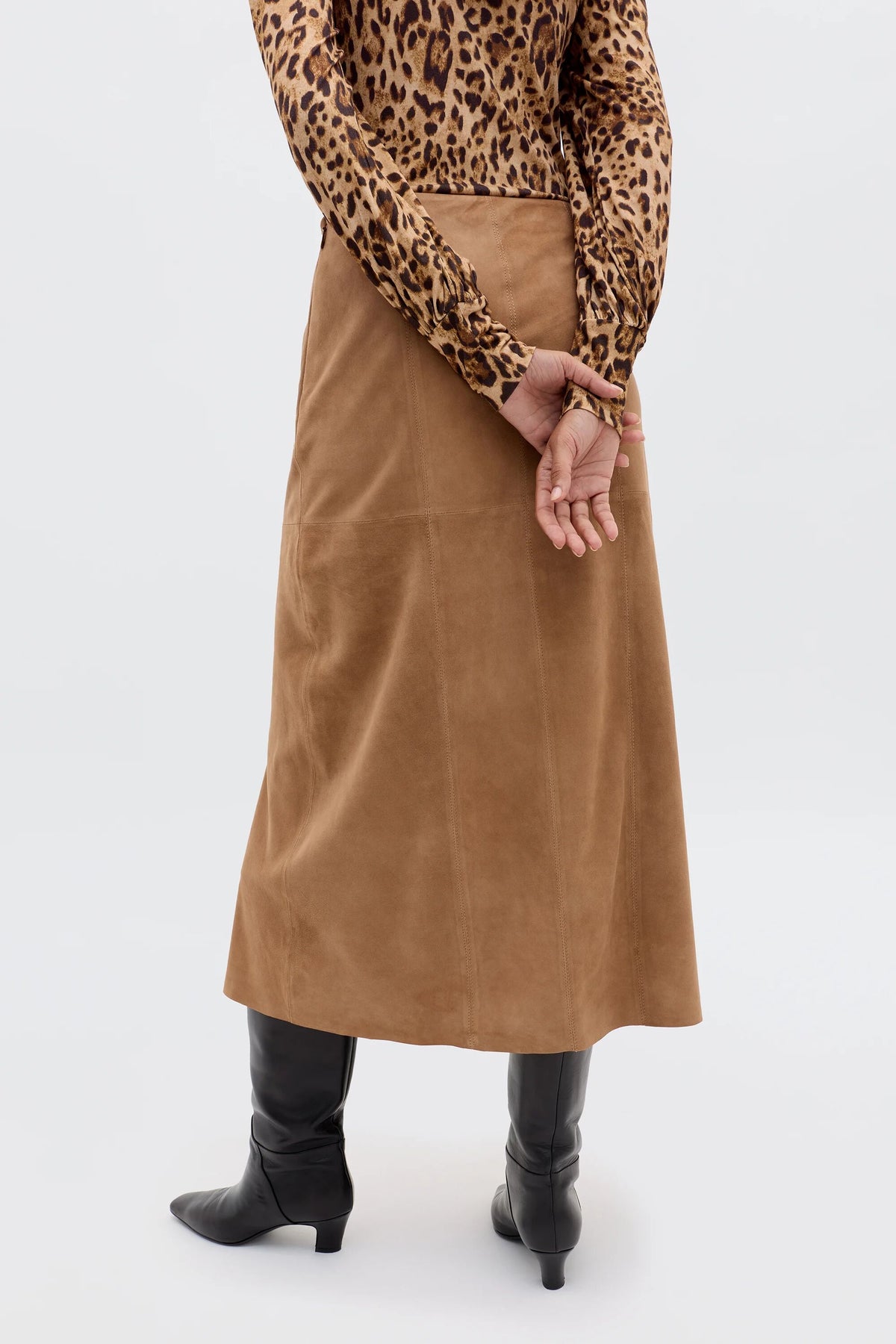 A person stands with their back to the camera, hands clasped behind them, wearing the Edwina - Camel high-waisted suede skirt, a leopard print long-sleeve top, and black knee-high heeled boots.