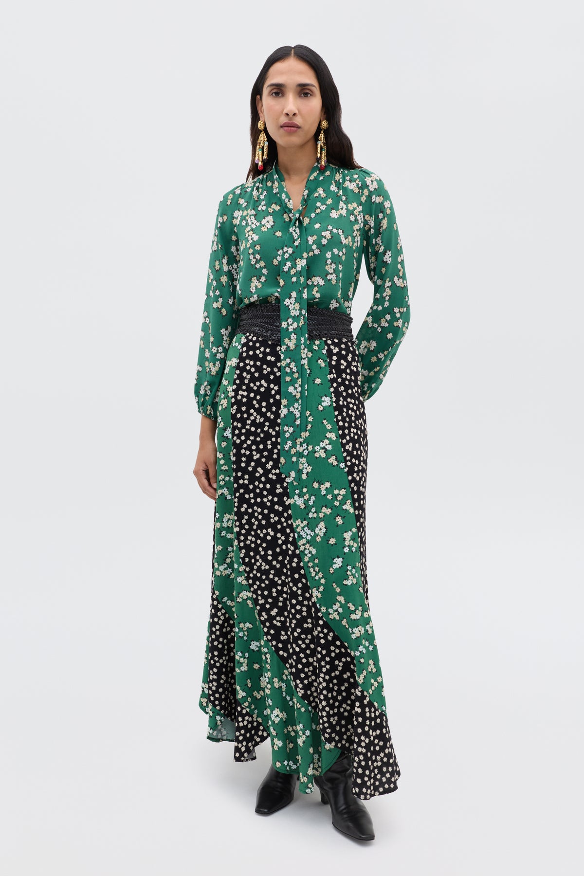 A woman with long dark hair wears the Gracella - City Floral Mix Olive maxi dress, featuring green and black florals, long sleeves, a belted waist, and black boots. Statement earrings complete her transitional look against a plain background.