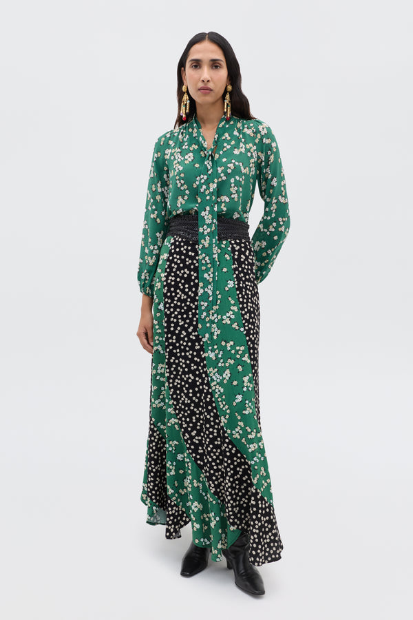A woman with long dark hair wears the Gracella - City Floral Mix Olive maxi dress, featuring green and black florals, long sleeves, a belted waist, and black boots. Statement earrings complete her transitional look against a plain background.