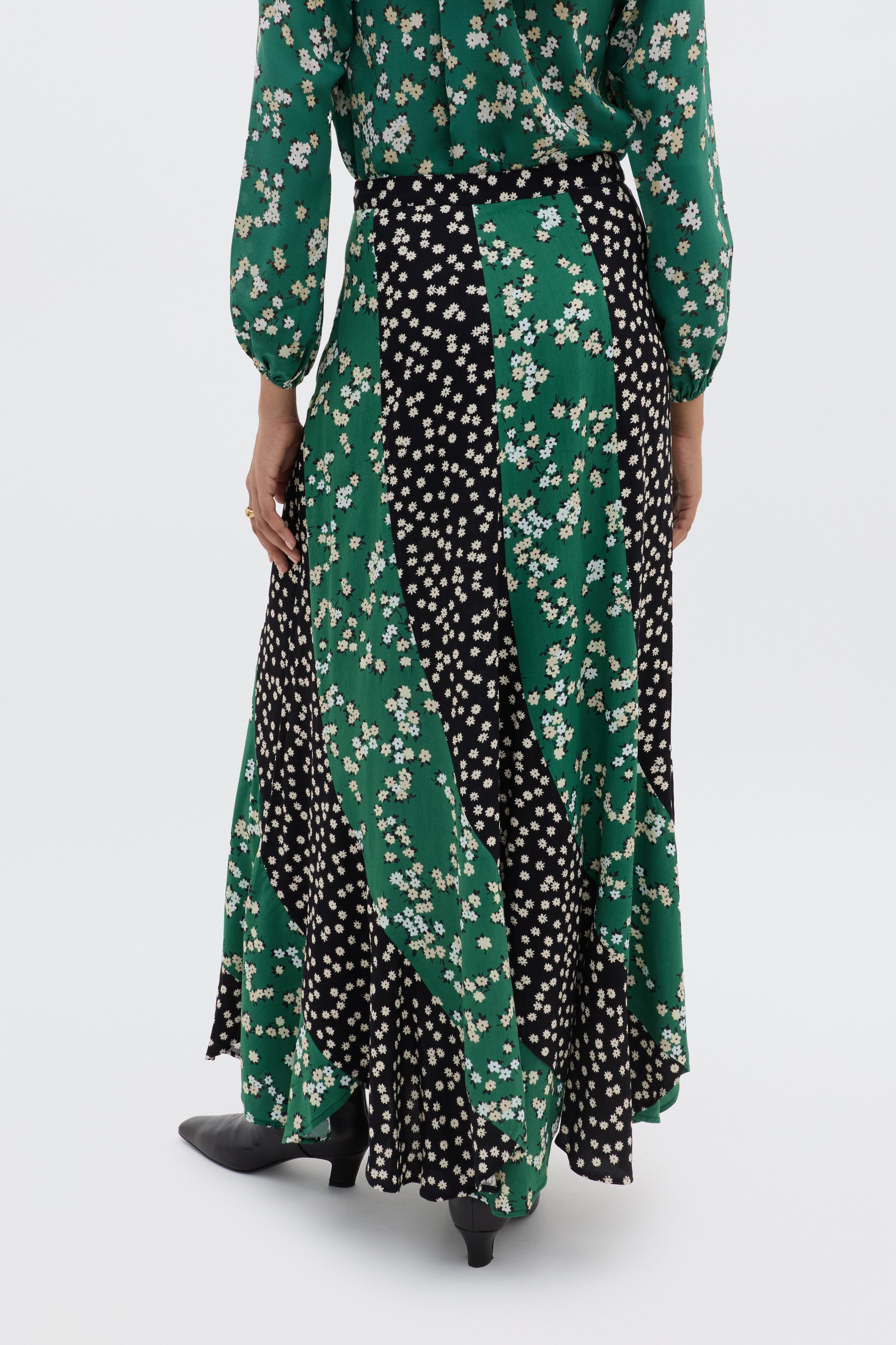 A person is shown from the back wearing the Gracella - City Floral Mix Olive, a long flowy midi skirt with alternating black and green panels featuring white winter floral prints, paired with black heeled shoes.
