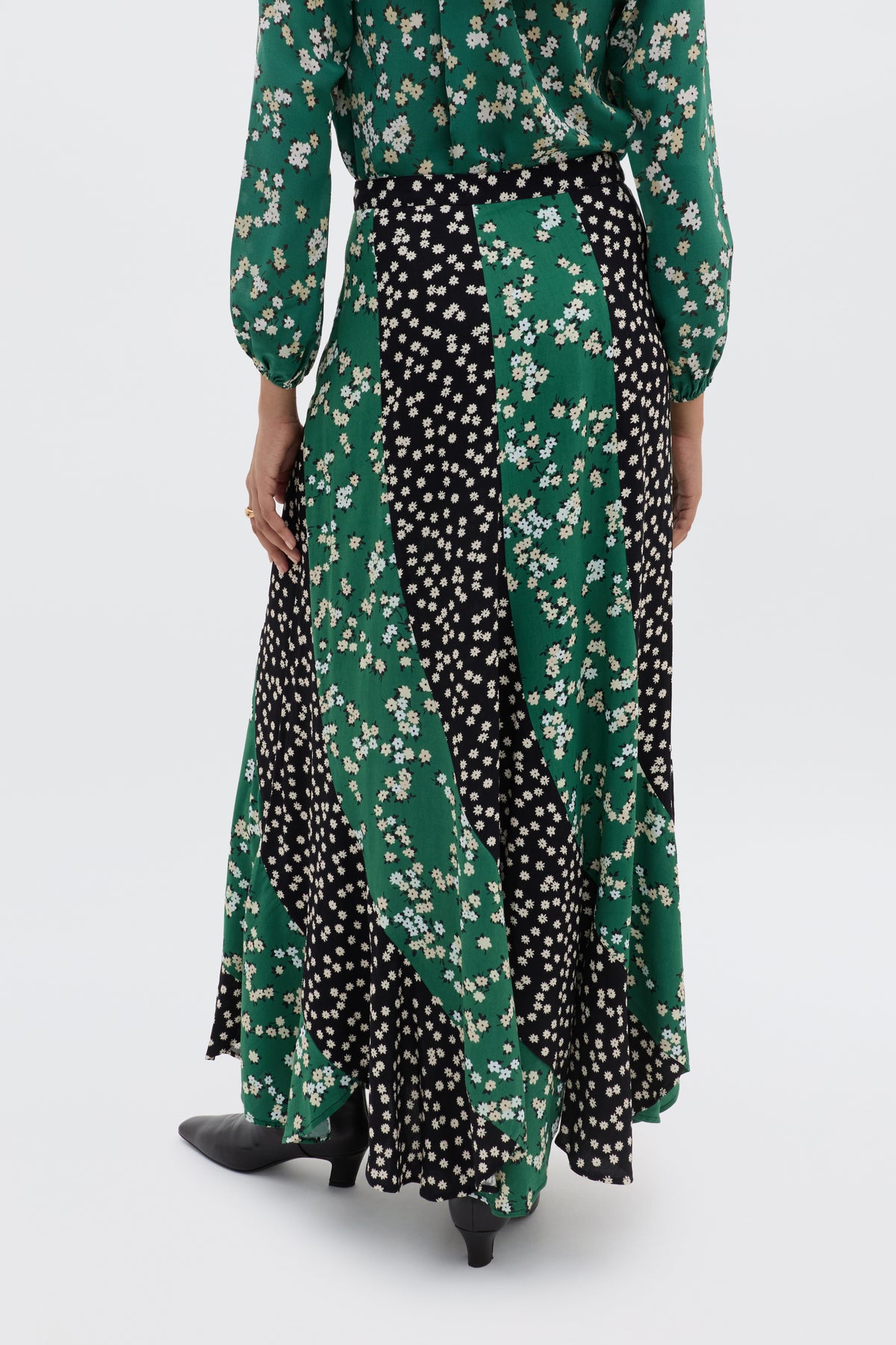 A person is shown from the back wearing the Gracella - City Floral Mix Olive, a long flowy midi skirt with alternating black and green panels featuring white winter floral prints, paired with black heeled shoes.