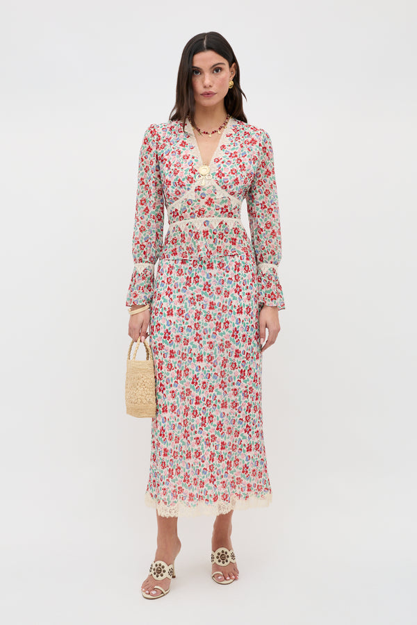 A woman stands against a plain white background wearing the Stassie - Jeannie Bloom Ivory: a long-sleeve, floral print midi dress with lace trim, paired with white sandals, layered necklaces, and a small woven handbag.