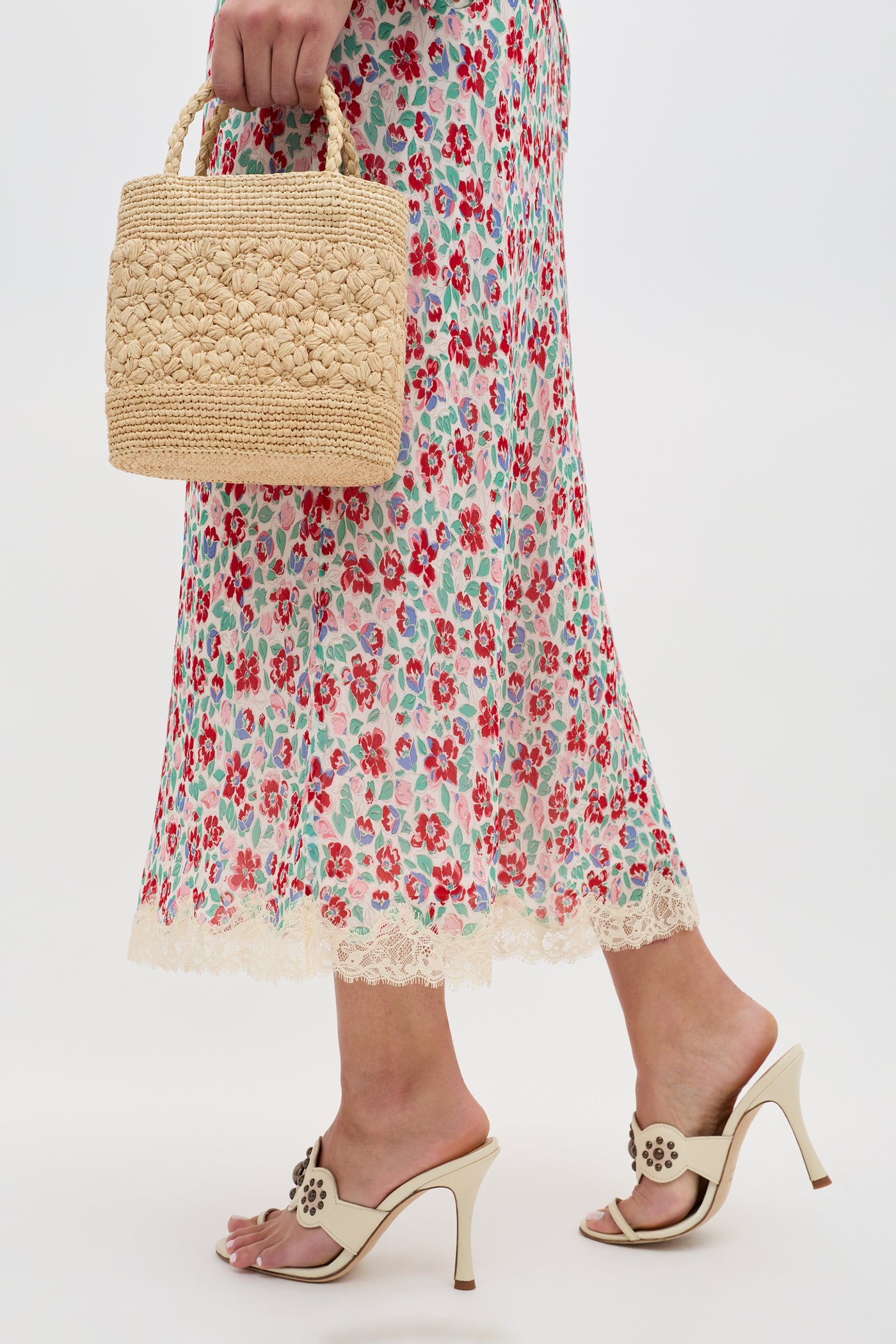A person holds the Stassie - Jeannie Bloom Ivory, a floral, lace-trimmed skirt, and pairs it with cream heeled sandals and a woven straw handbag against a plain background.