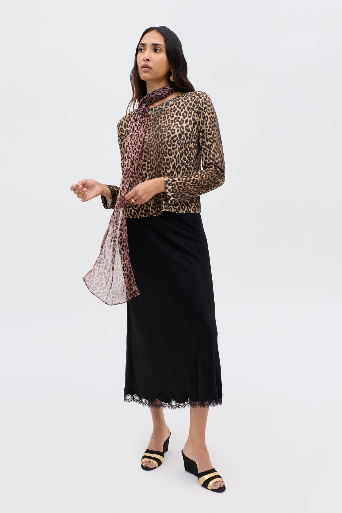 In Paris, a woman stands against a plain background, wearing the Stassie - Black sandals with a leopard-print top, sheer matching scarf, and a black satin skirt featuring wavy lace trim.