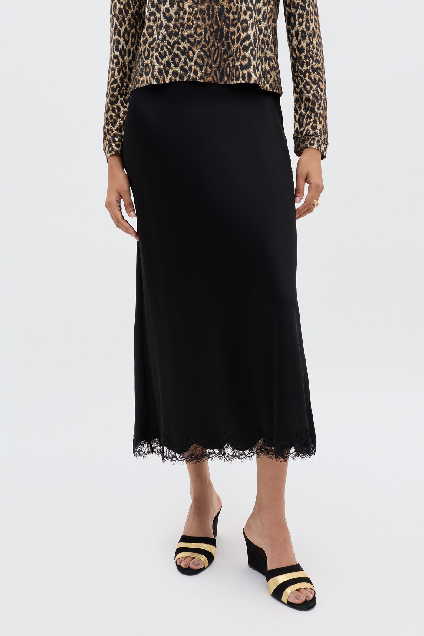 Wearing the Stassie - Black heeled sandals, a leopard print long-sleeve top, and a wavy lace-trimmed satin skirt, she stands before a white backdrop—effortlessly embodying Parisian style.