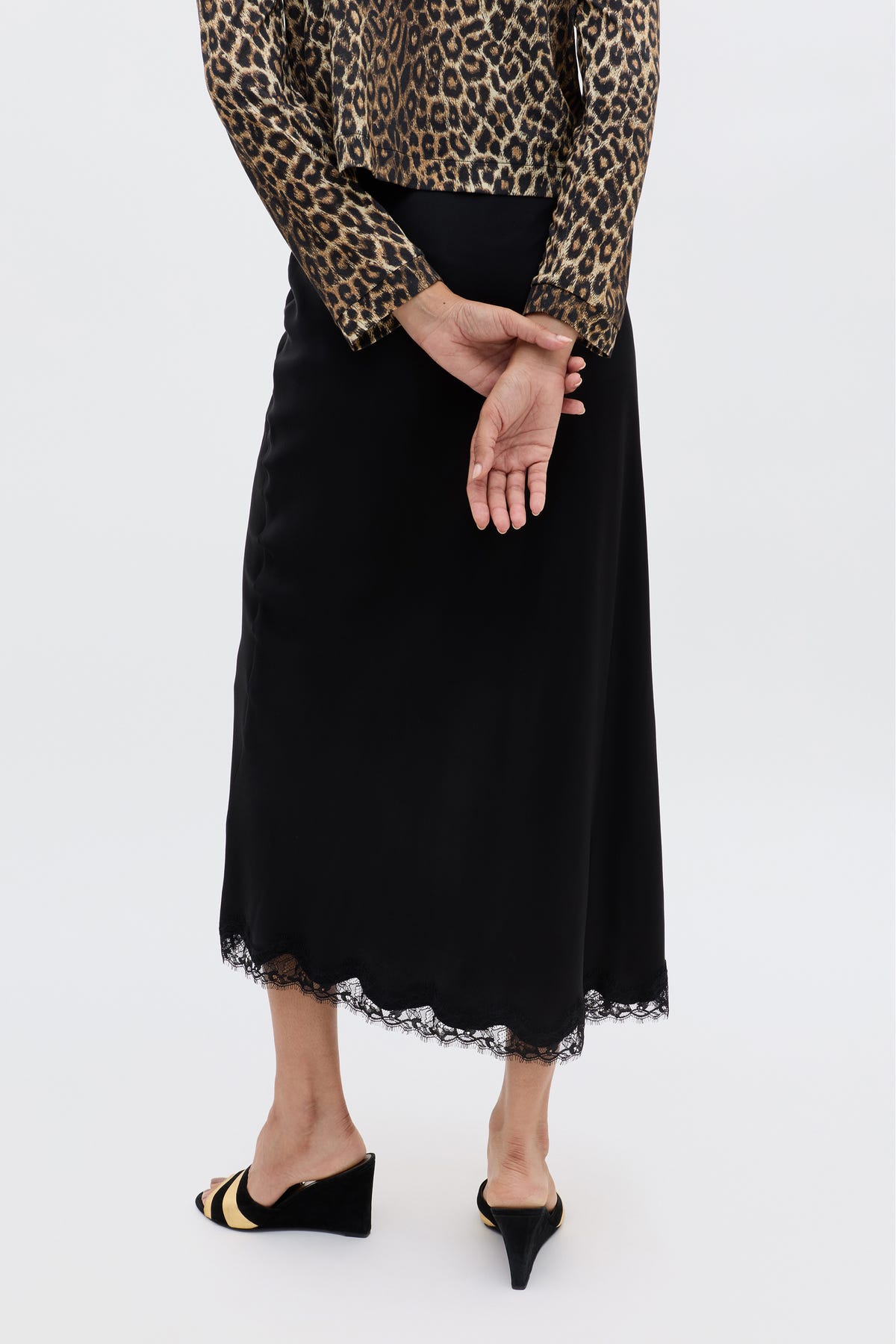 Clad in the Stassie - Black skirt with wavy lace trim, a leopard print long-sleeve top, and black wedge sandals, the person stands hands clasped behind their back, channeling effortless Parisian style against a plain backdrop.