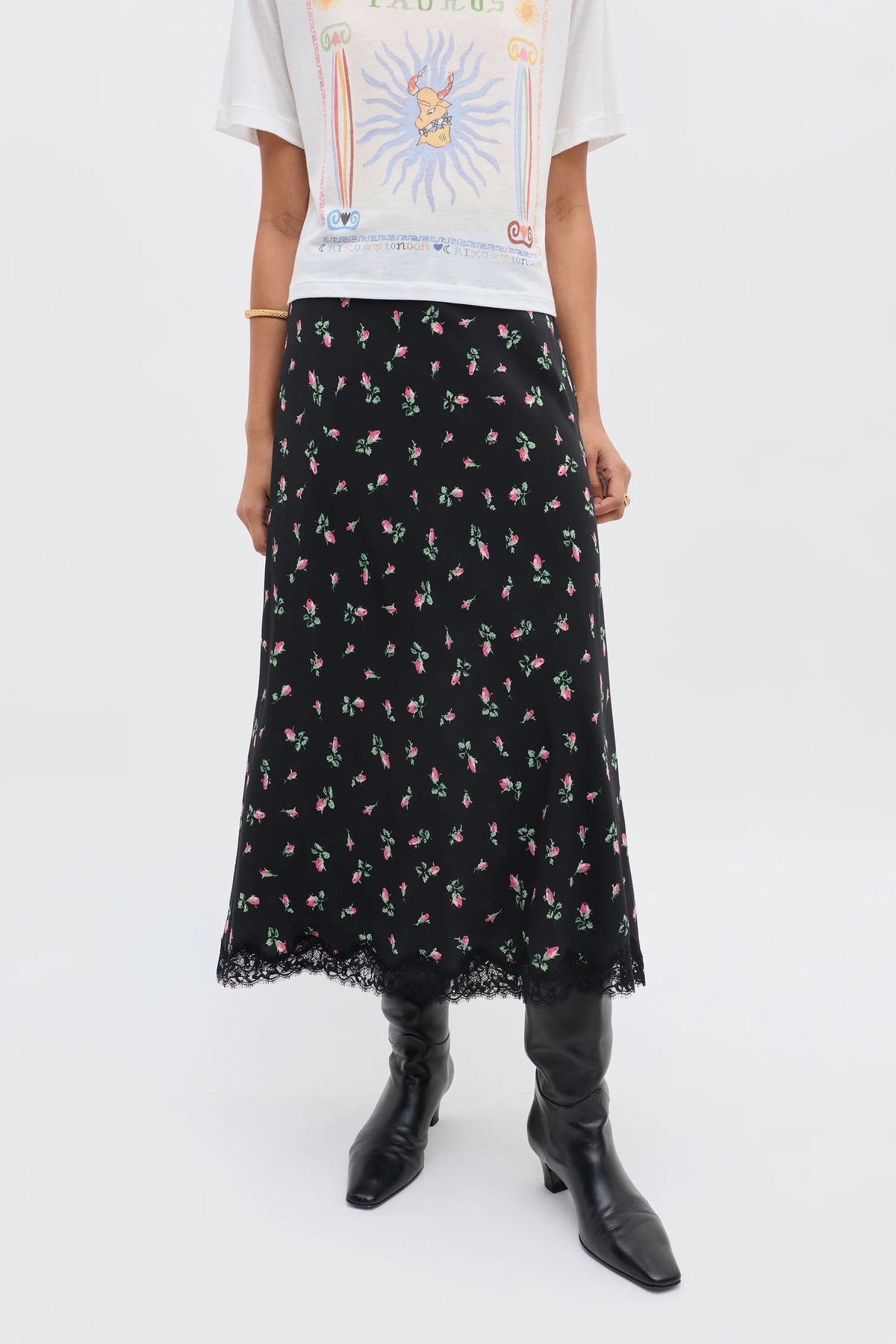 A person wears the Stassie - Sienna Bud Black vintage-inspired black midi skirt with a small pink floral print and lace hem, paired with a white graphic t-shirt and black heeled boots. The photo is cropped at the shoulders.