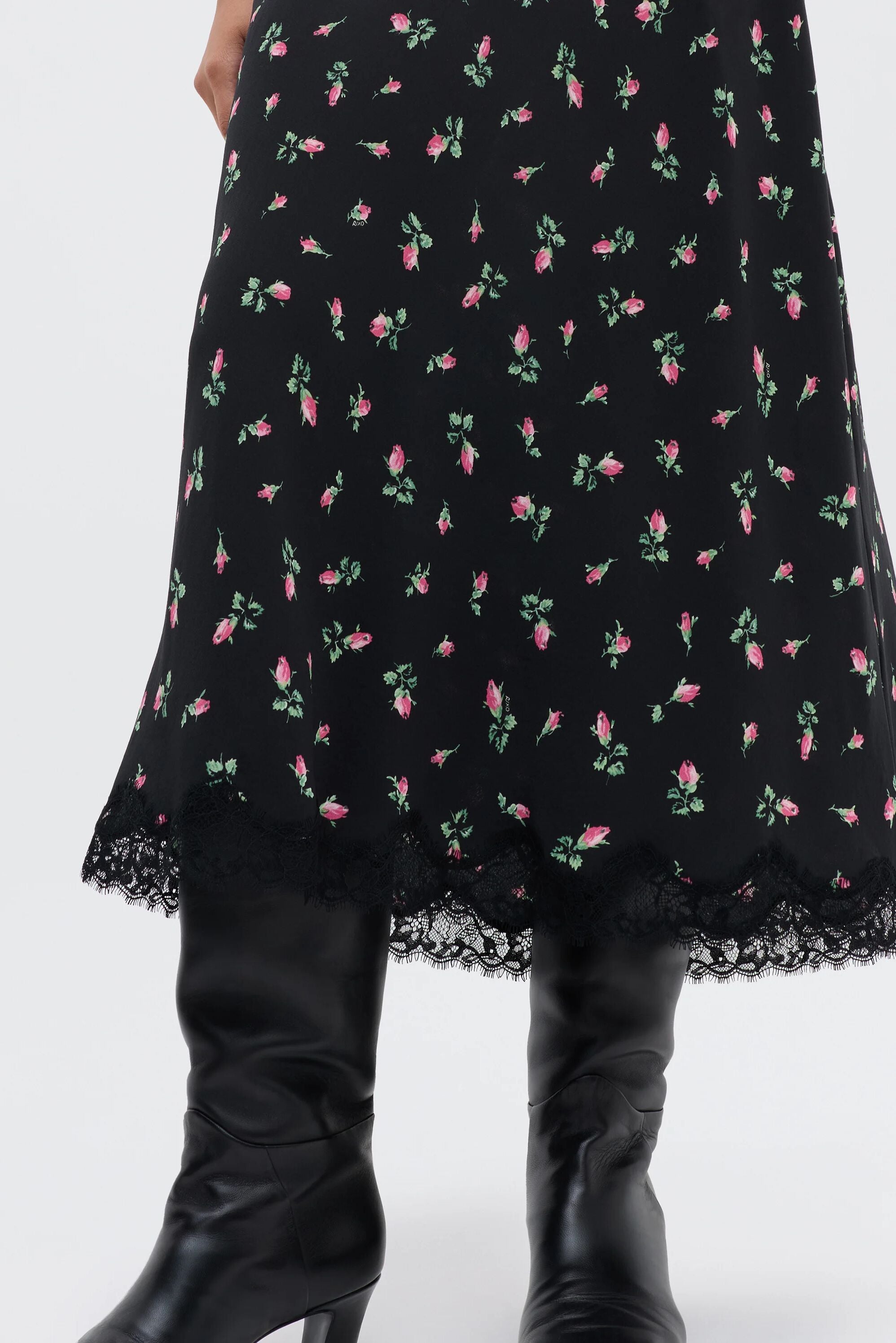 A person in the Stassie - Sienna Bud Black skirt, featuring a pink rose floral pattern and lace hem, paired with tall black boots, stands against a plain light background.