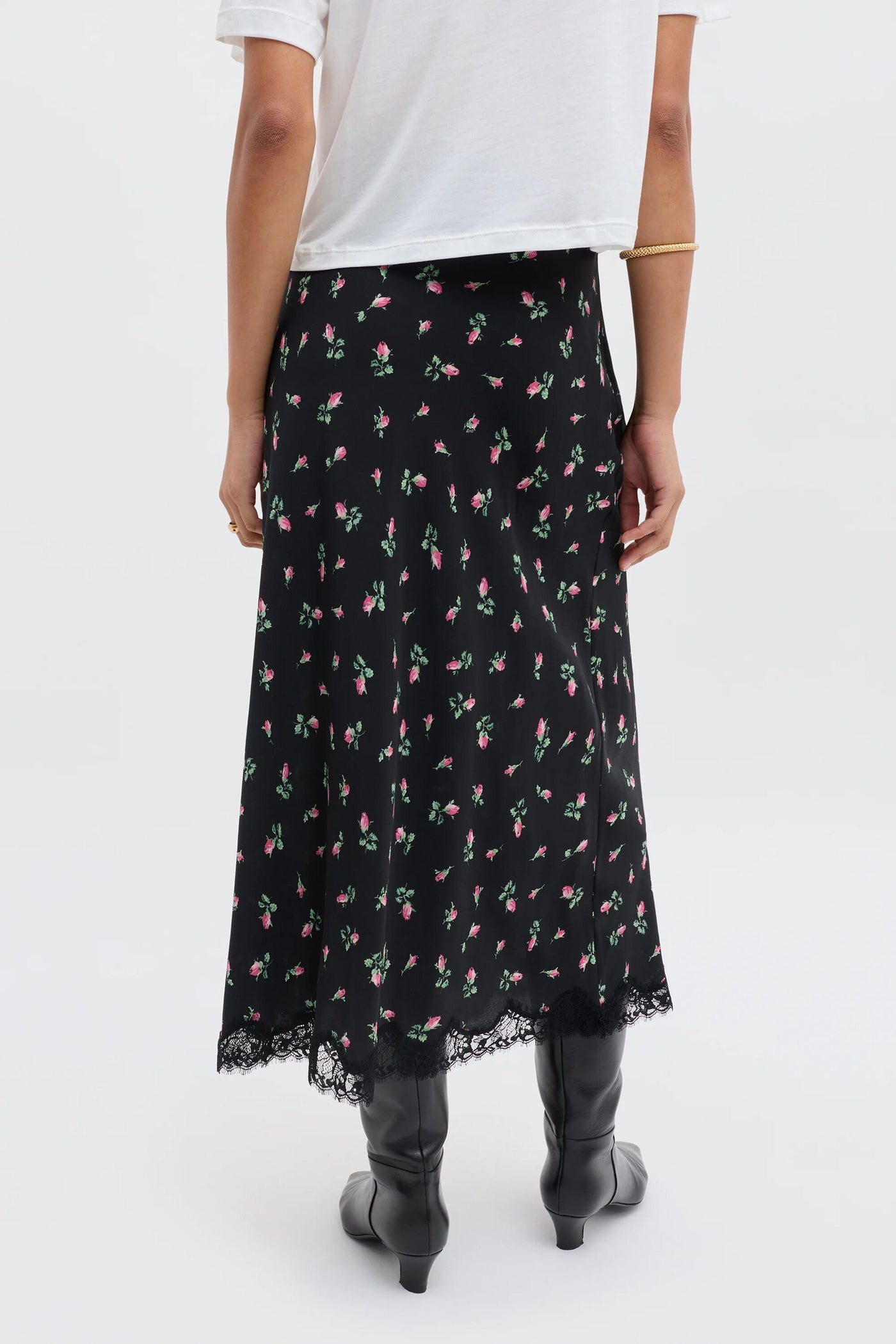 A person is shown from the back wearing the Stassie - Sienna Bud Black skirt, featuring a black midi length with a small pink floral pattern and lace hem, paired with a white short-sleeve shirt and black knee-high boots.