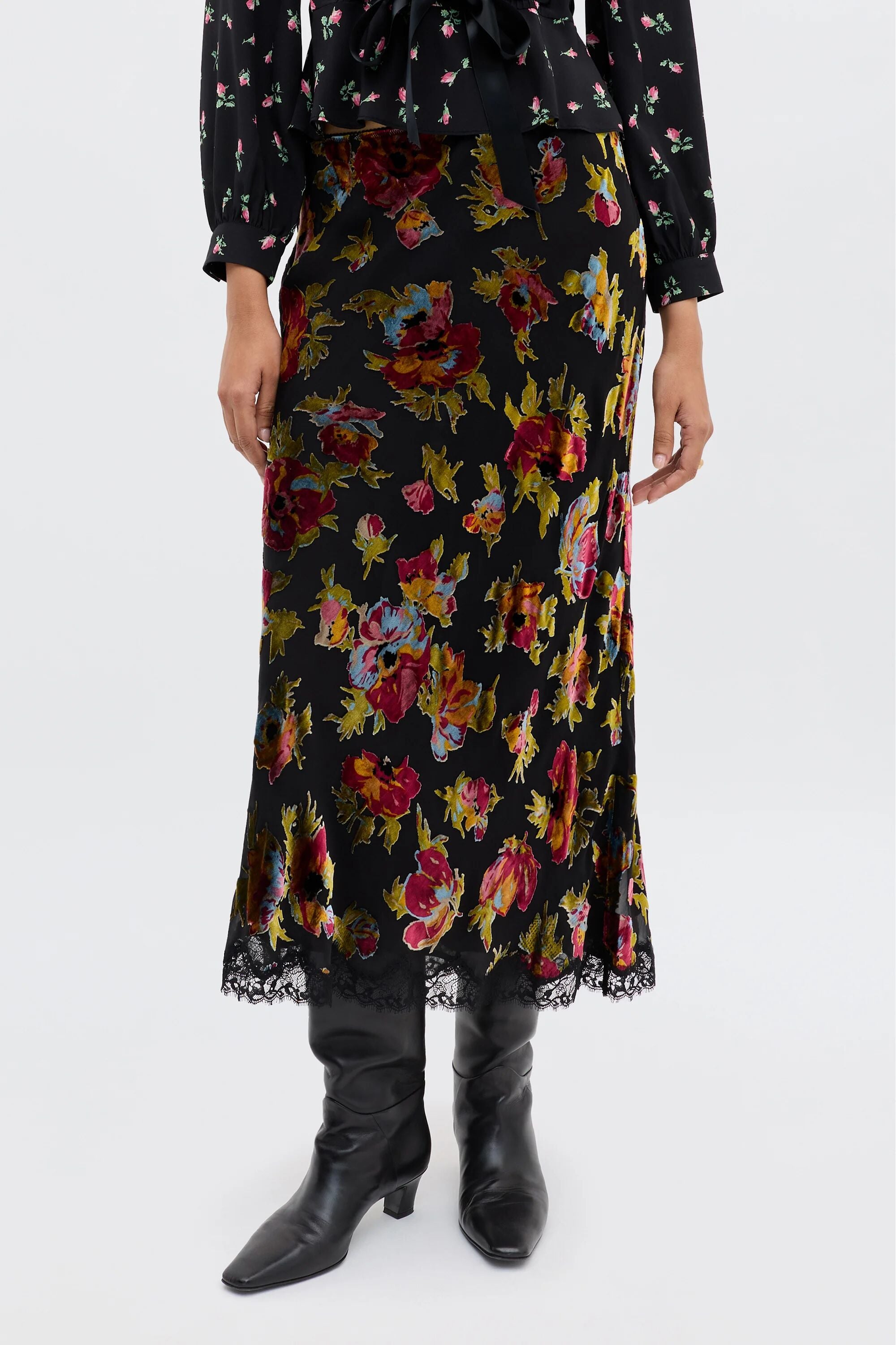 A person wearing the Stassie - Countryside Devoré Black midi skirt featuring a colorful floral pattern, lace hem trim, and black knee-high boots. The top of a matching black floral blouse is partially visible.