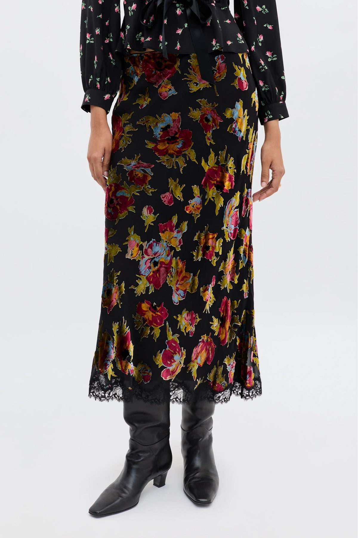 A person wearing the Stassie - Countryside Devoré Black midi skirt featuring a colorful floral pattern, lace hem trim, and black knee-high boots. The top of a matching black floral blouse is partially visible.