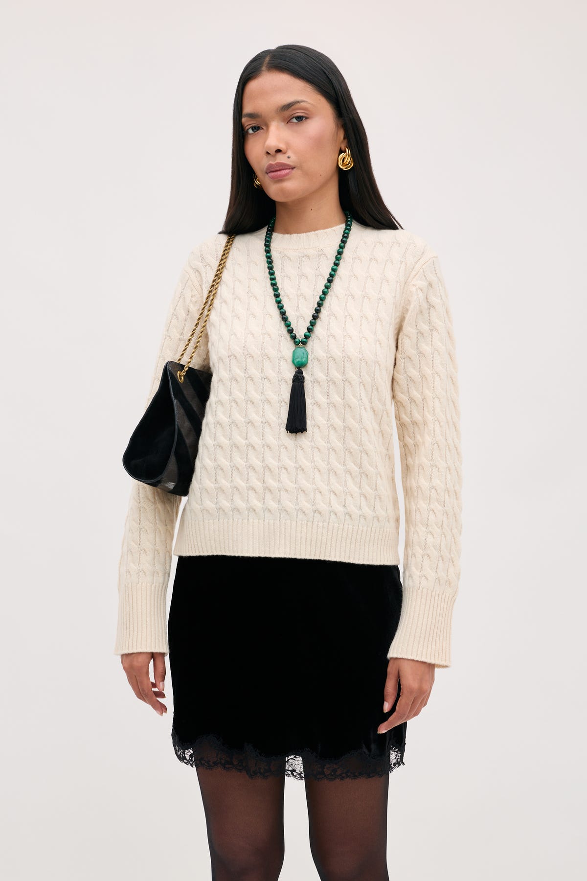 A woman wears a cream cable-knit sweater, a Kornelia - Black velvet mini skirt with a lace hem, and a black shoulder bag with a gold chain strap.