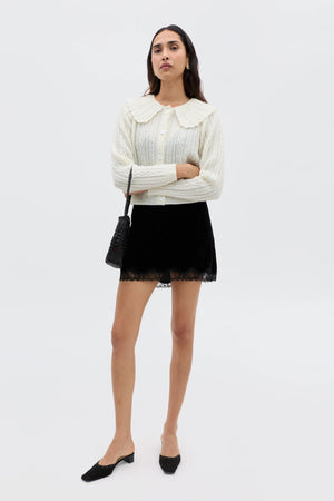 A woman poses against a white background, wearing a cream cable-knit cardigan, a lace-trimmed velvet mini skirt for '90s flair, black flats, and carrying Kornelia - Black, a small black shoulder bag.