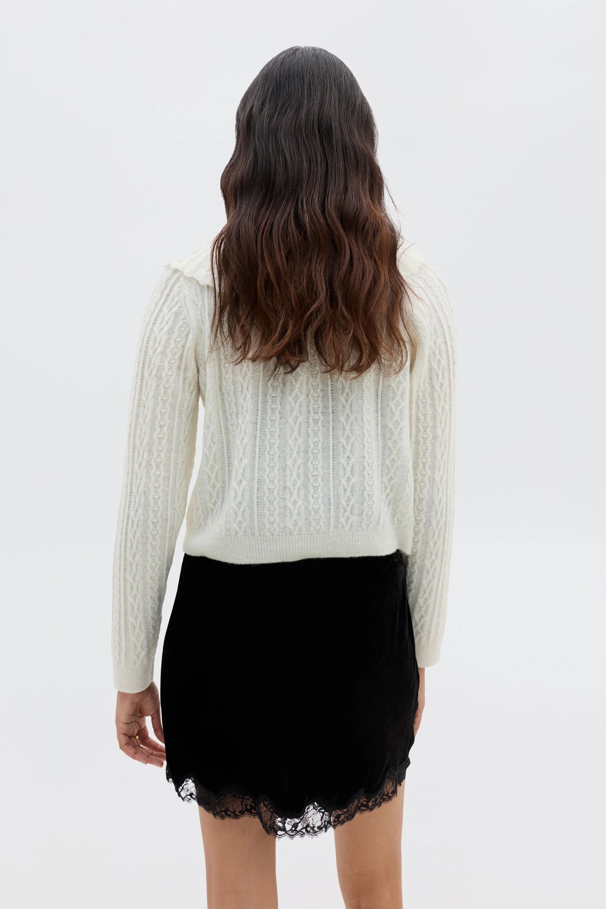 A person with wavy brown hair, seen from behind, models Kornelia - Black: a nineties style black velvet mini skirt with lace trim and a cream knit sweater, against a plain white background.
