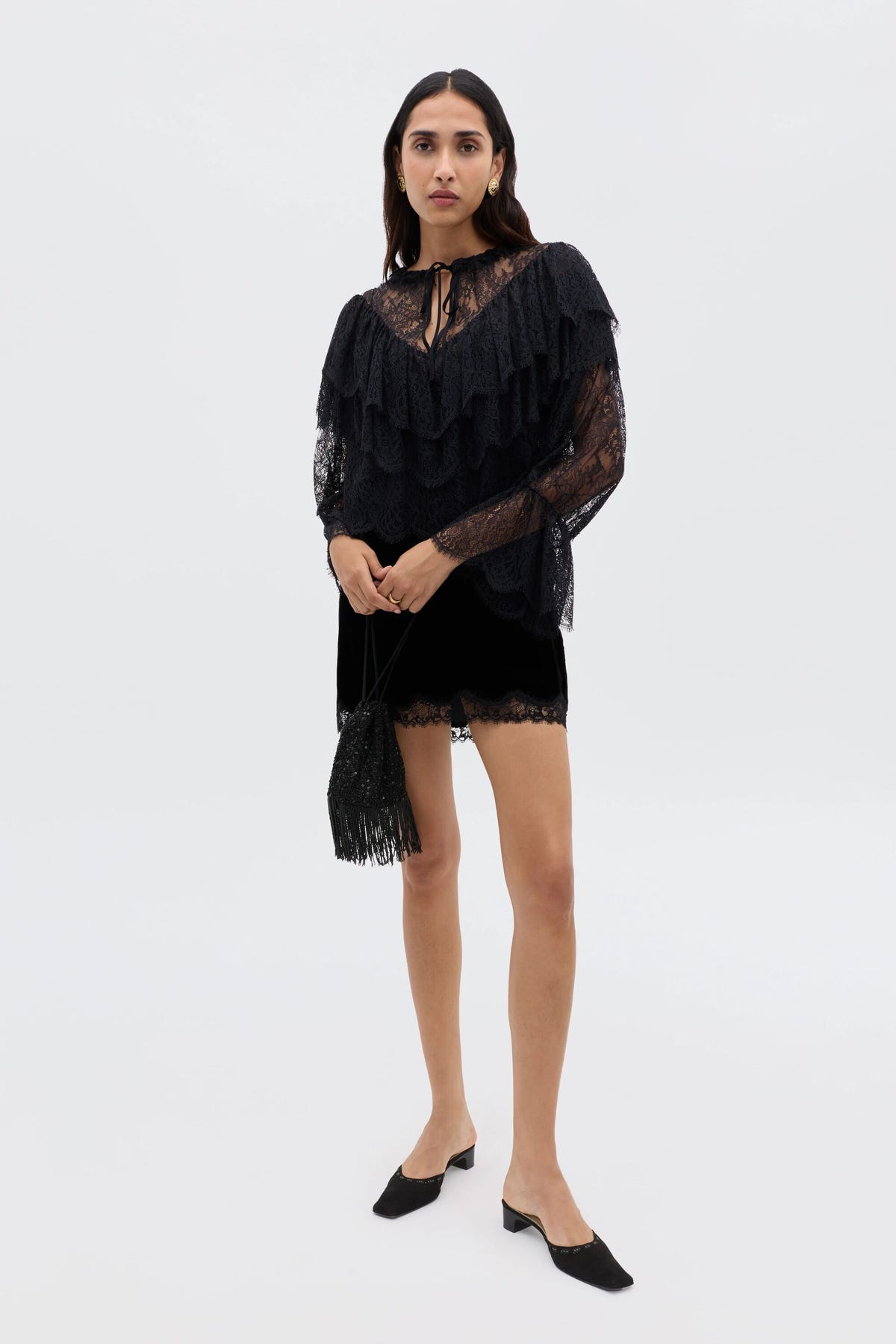 Against a white background, a woman wears the Kornelia - Black: a black lace top with ruffles, velvet mini skirt, mule shoes, and carries a small fringed black handbag for Nineties-inspired flair.