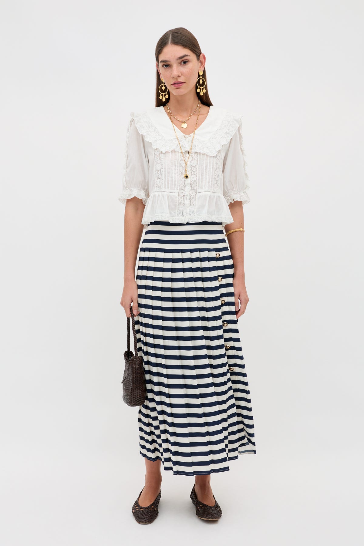 A woman stands against a white background wearing the Una - Lea Stripe Navy skirt, a white blouse with a large collar, black flats, gold statement earrings, layered necklaces, and holding a small black bag.