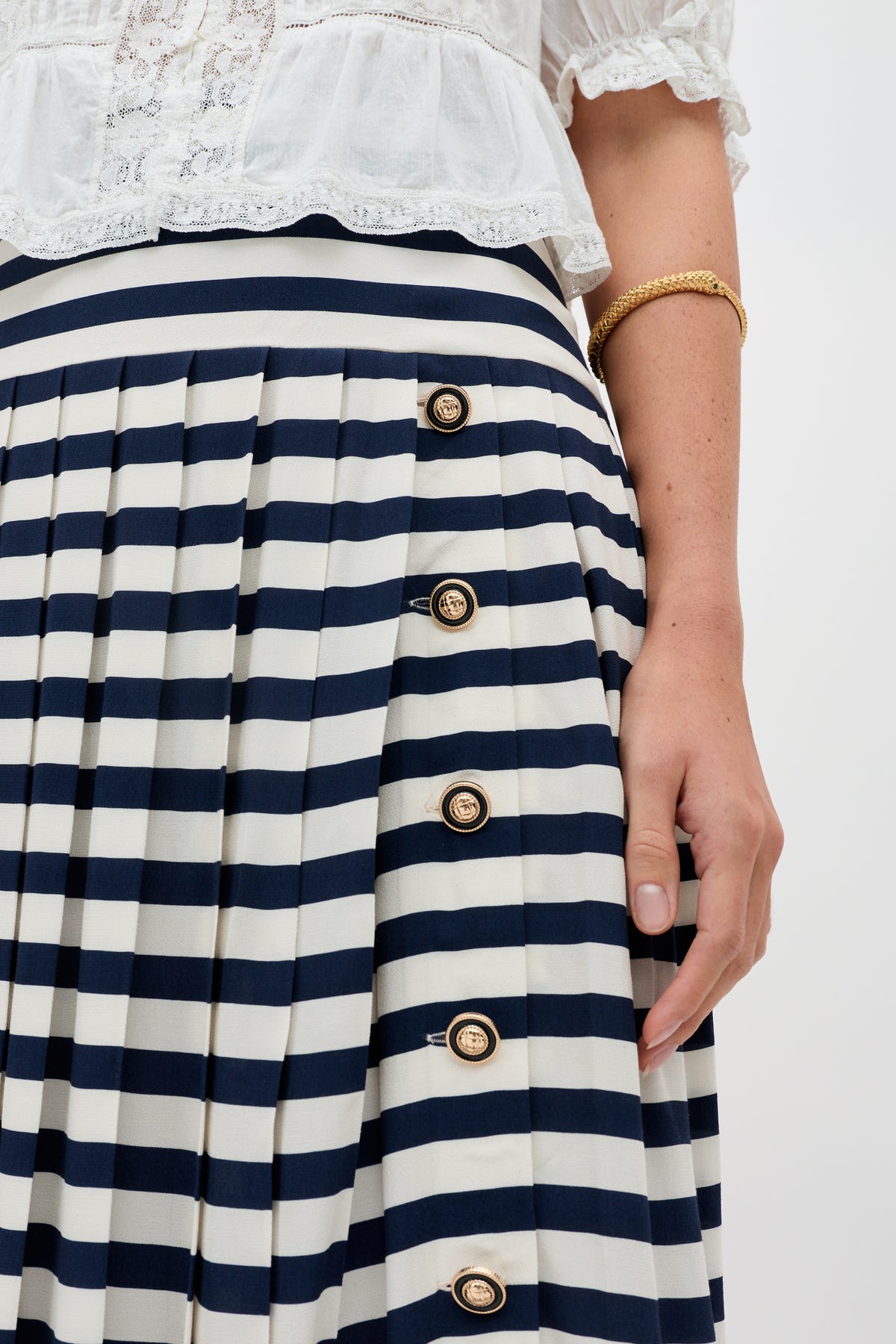 Wearing the Una - Lea Stripe Navy skirt, featuring pleats and gold buttons, paired with a white blouse and a gold bangle. Only the lower torso and hand are shown.