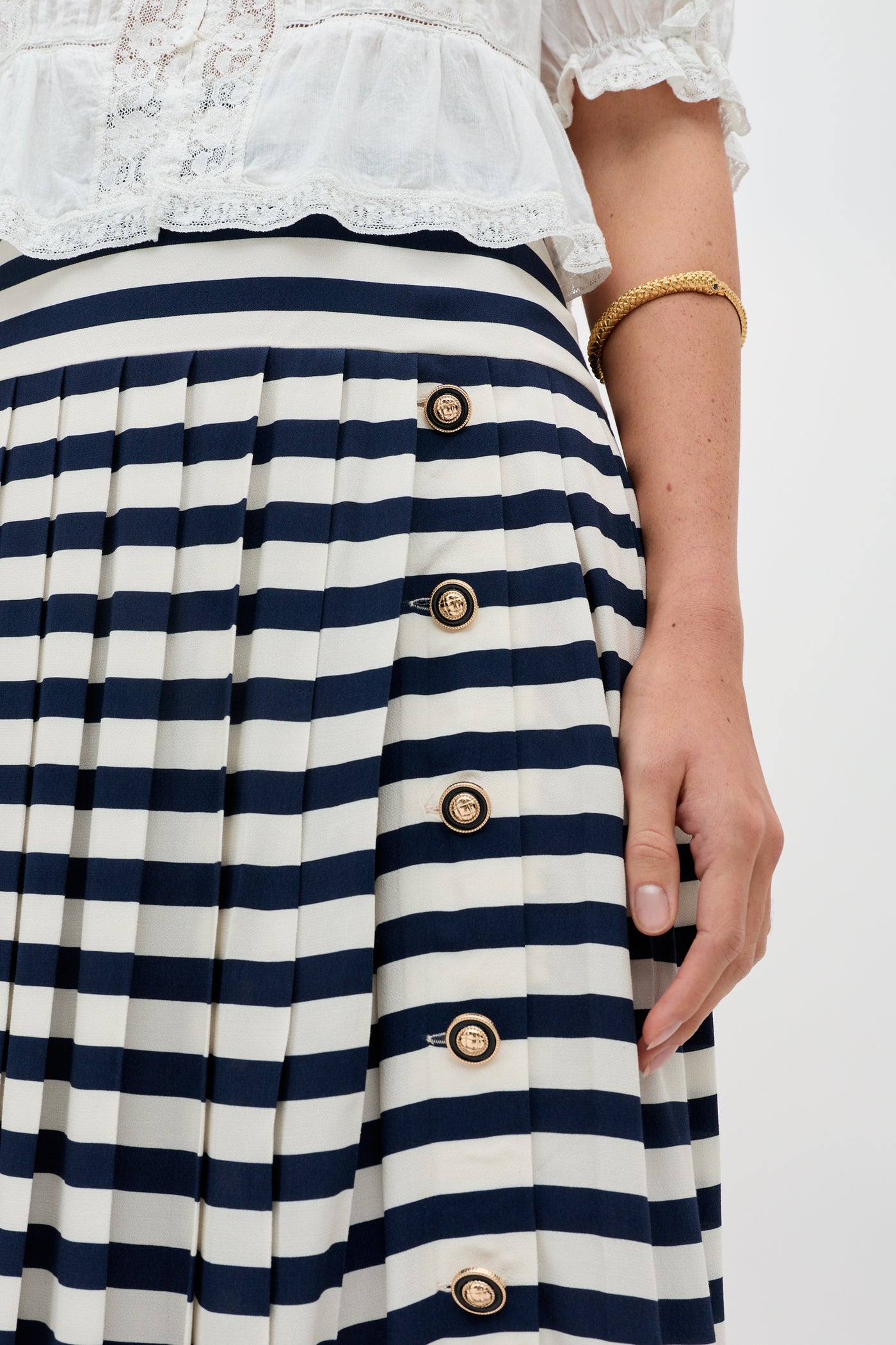 Wearing the Una - Lea Stripe Navy skirt, featuring pleats and gold buttons, paired with a white blouse and a gold bangle. Only the lower torso and hand are shown.