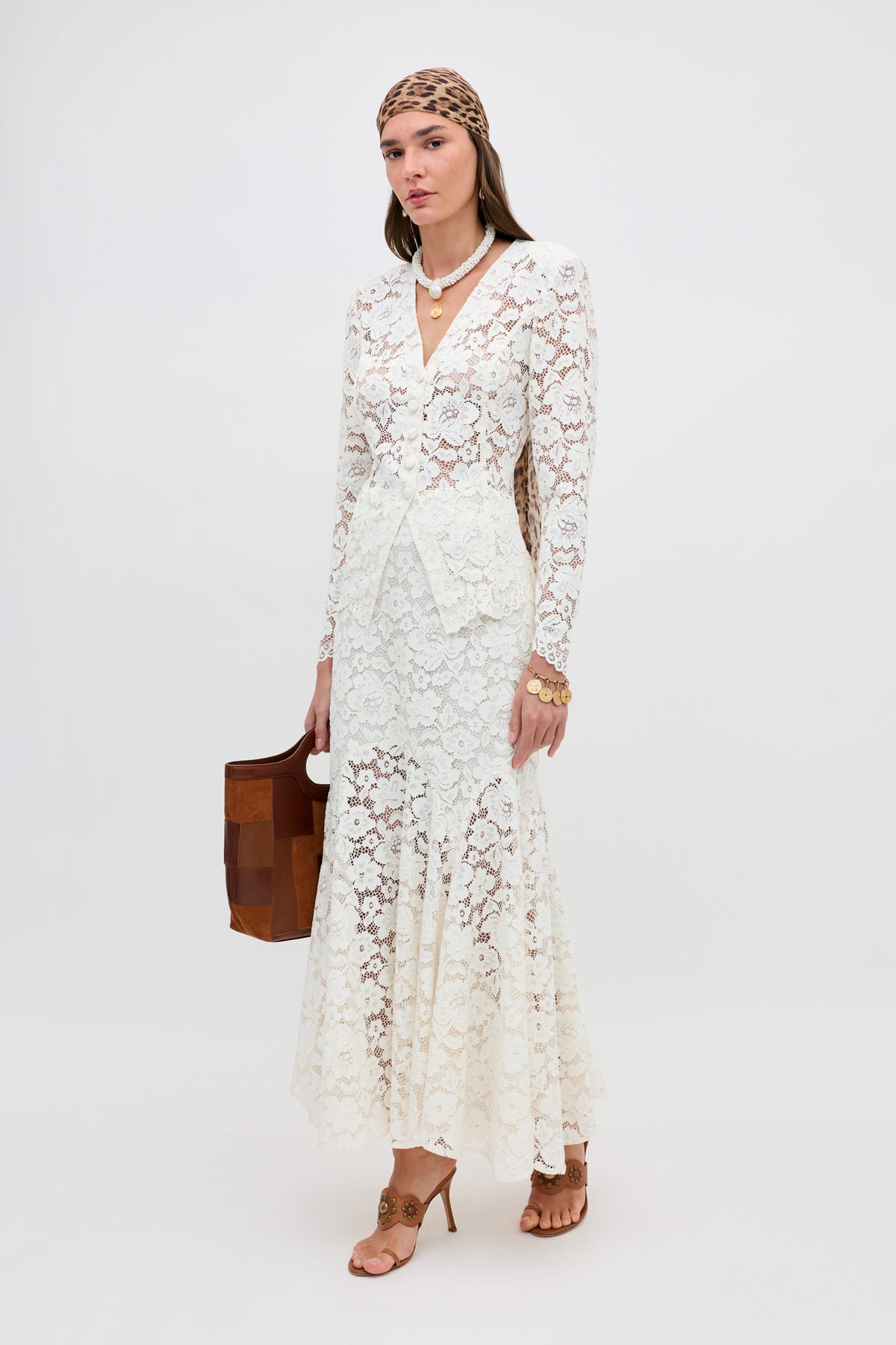 A woman stands against a plain background wearing the Jada - Cream long lace dress with long sleeves, tan heels, a brown patterned headscarf, layered necklaces, gold jewelry, and carrying a brown handbag.