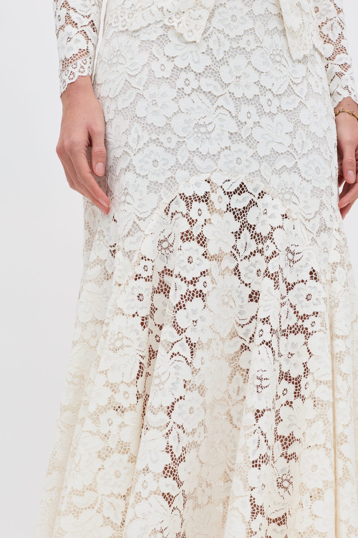 A close-up of a person wearing the Jada - Cream dress, featuring long white lace with floral patterns, shown from the waist to the knees and one hand resting at their side.