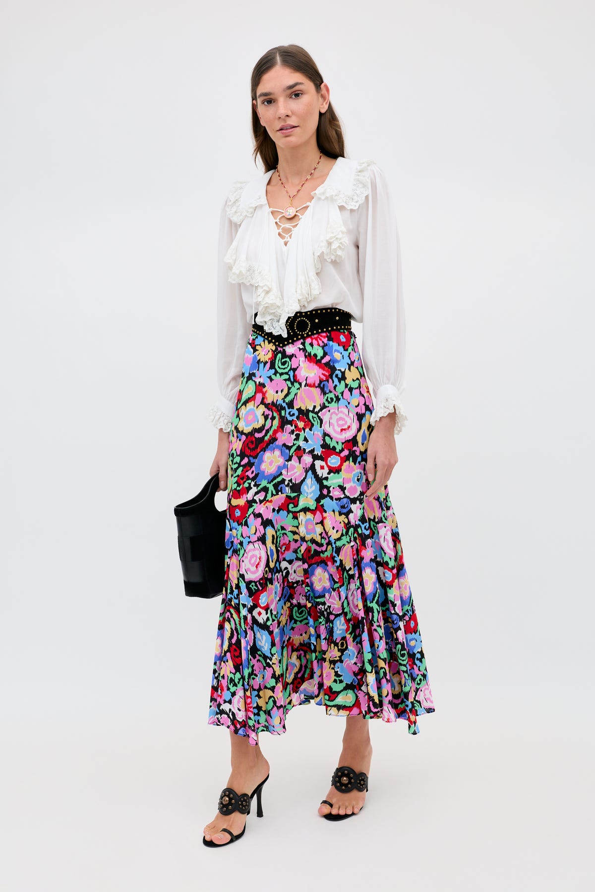 A woman poses against a plain background wearing the Libbie - Ikat Bloom Charcoal floral midi skirt, a white ruffled blouse, black sandals with a matching belt, layered necklaces, and carrying a black handbag.