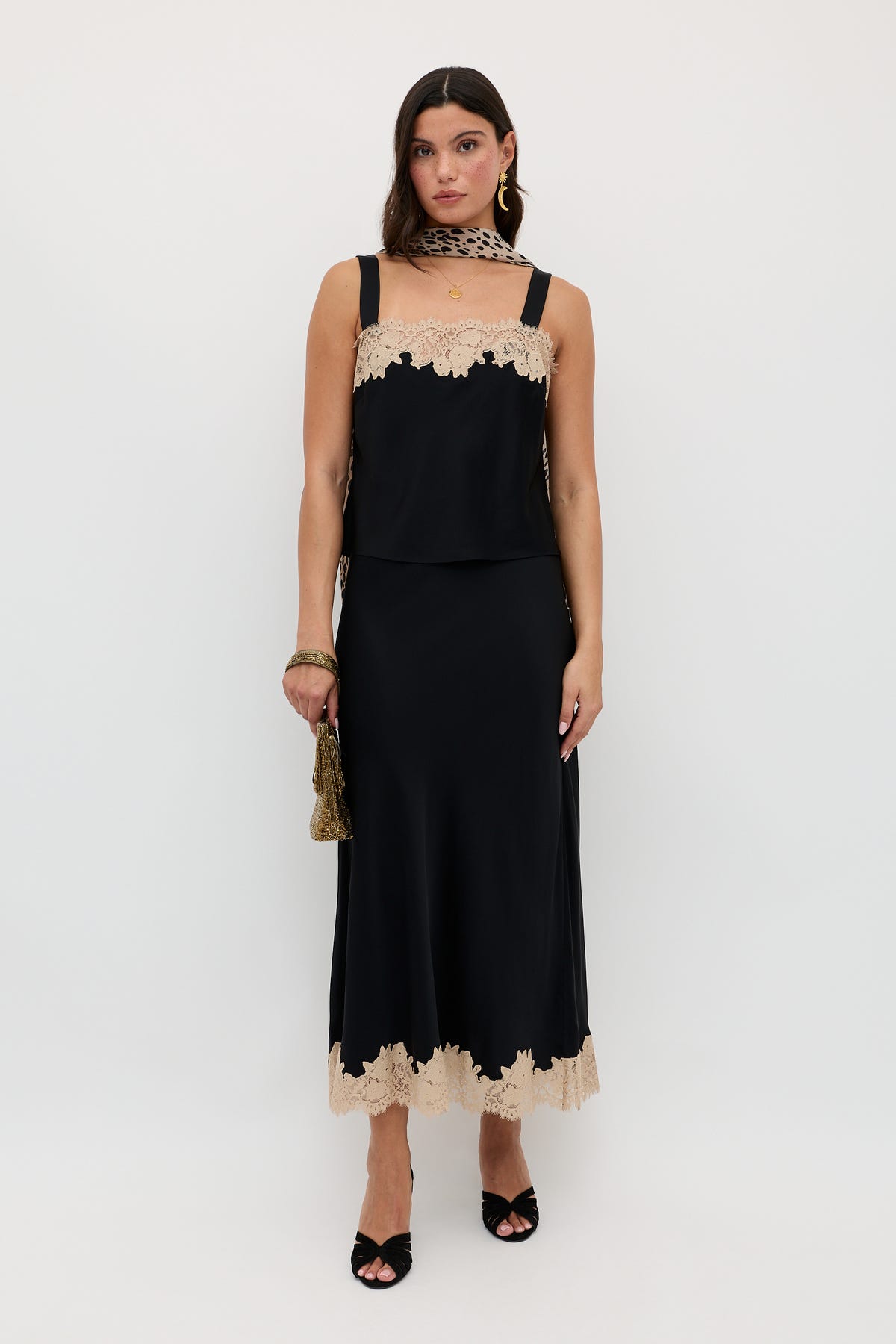 A woman wearing the Matilde - Black sleeveless dress with beige lace trim at the neckline and hem, accessorized with a small gold bag, black heels, and a beige lace scarf, stands against a plain white background.