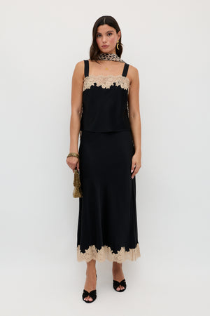 A woman wearing the Matilde - Black sleeveless dress with beige lace trim at the neckline and hem, accessorized with a small gold bag, black heels, and a beige lace scarf, stands against a plain white background.