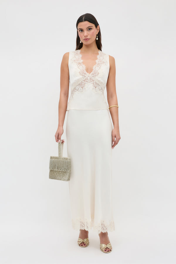 A woman models the Matilde - Pearl dress, a sleeveless cream gown with lace at the neckline and hem. She pairs it with a textured beige handbag, drop earrings, a bangle, and heeled sandals against a white backdrop.