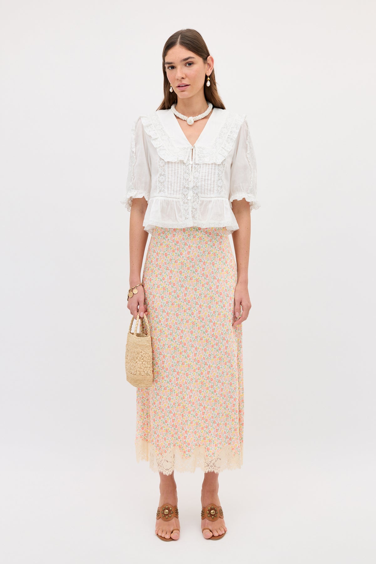 A woman stands against a plain background wearing a white blouse with lace collar, brown sandals, pearl jewelry, a woven handbag, and the Matilde - Katie Floral Cream long slip skirt with lace detailing.