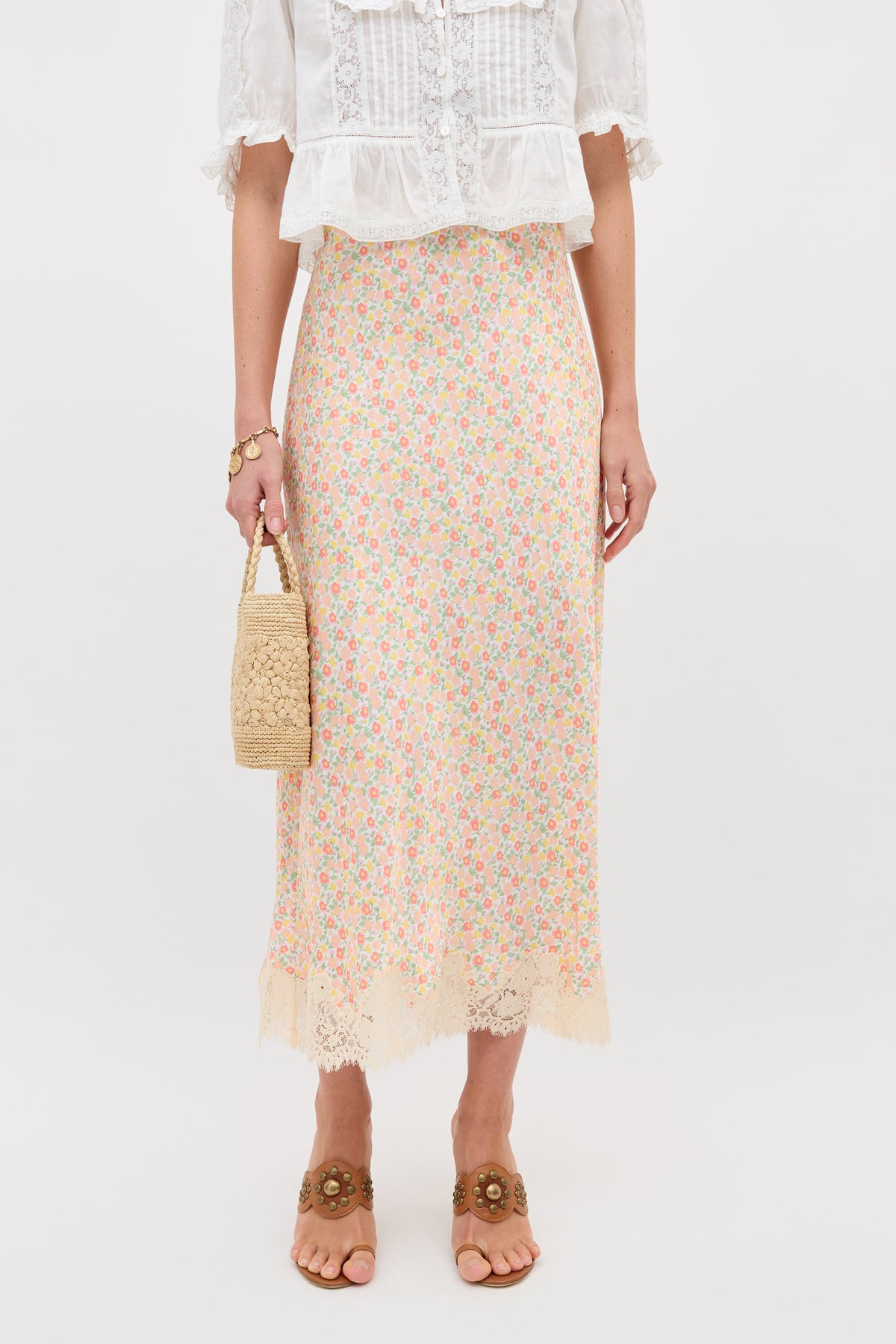 A woman wears the Matilde - Katie Floral Cream slip skirt with lace trim, a white lace blouse, brown floral sandals, and holds a woven straw bag. She stands against a plain white background.