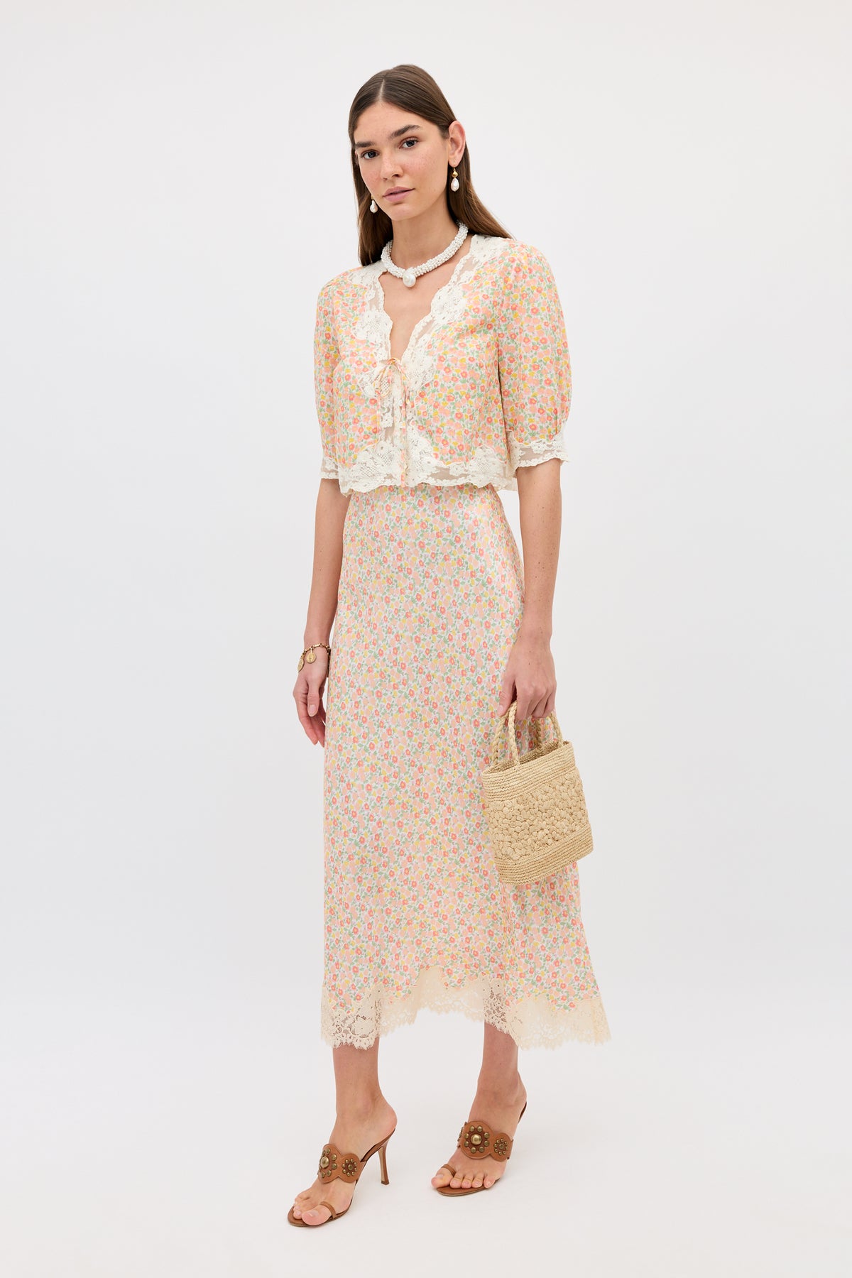 A woman stands against a white background wearing the Matilde - Katie Floral Cream two-piece with lace details, brown heeled sandals, pearl jewelry, and a small woven handbag. Her hair is down and she has a neutral expression.