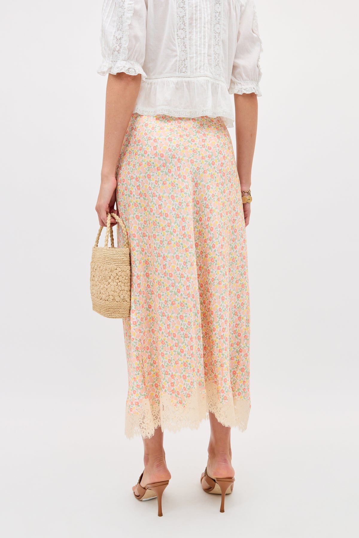 A woman faces away, wearing the Matilde - Katie Floral Cream skirt featuring pastel florals and lace at the hem, paired with a white lace-trim blouse, nude heels, and a small woven handbag against a plain light background.