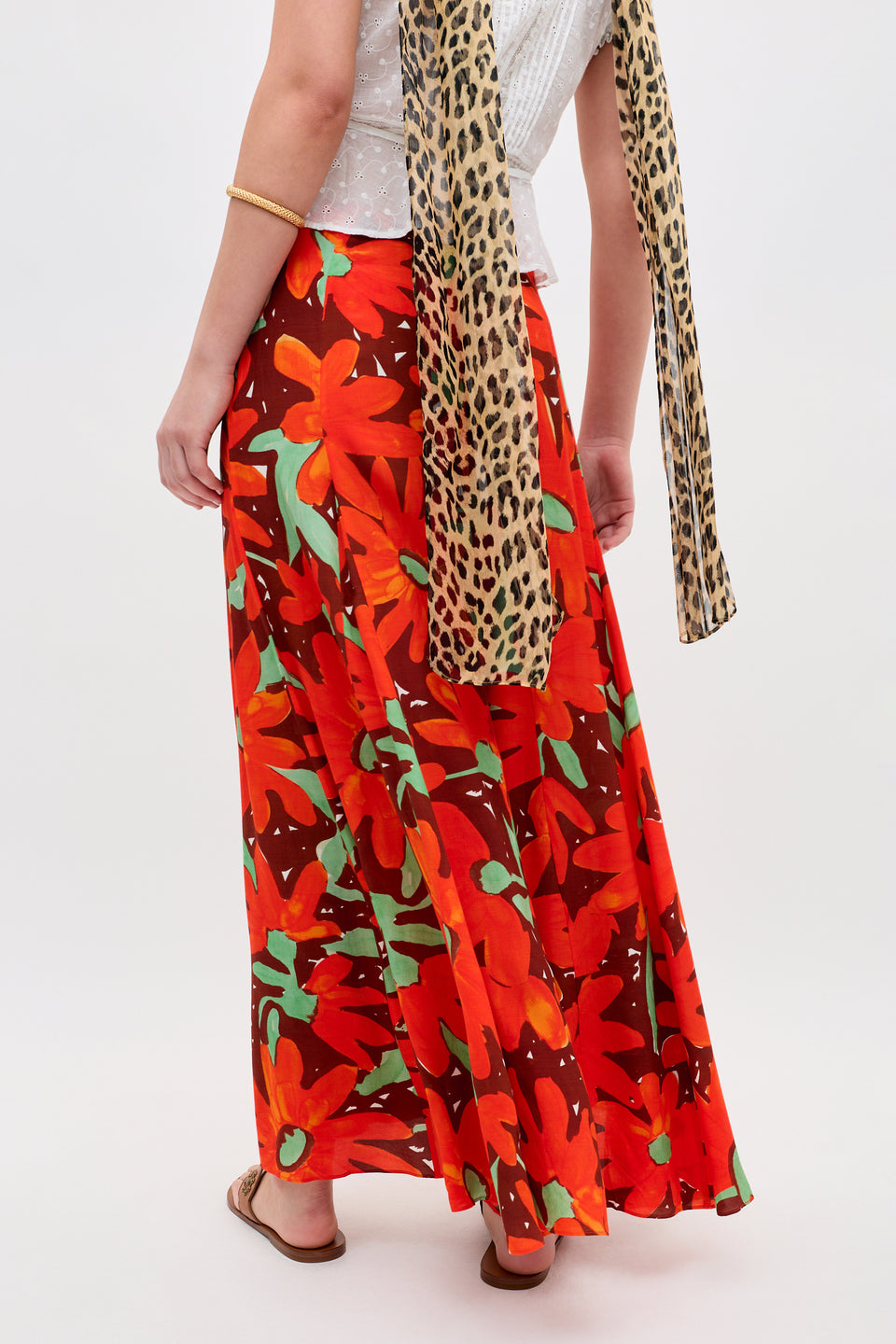 A person models Billy - Danish Floral Orange, a red maxi skirt with bold orange and green florals, paired with a white top, leopard print scarf, and sandals against a plain background.