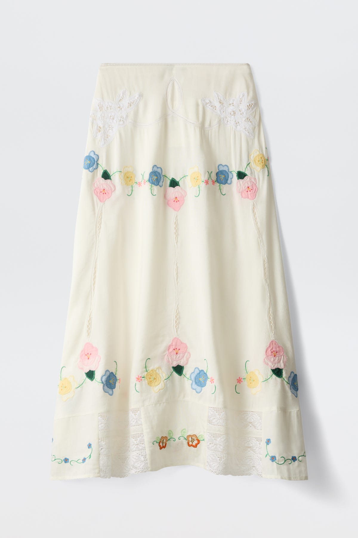 Paulina - Silje Embroidery Ivory is a long, cream-colored skirt adorned with delicate pink, blue, and yellow floral embroidery in pastel tones. Featuring lace details near the hem and decorative stitching along the length, this skirt showcases elegant embroidered flowers and intricate accents.