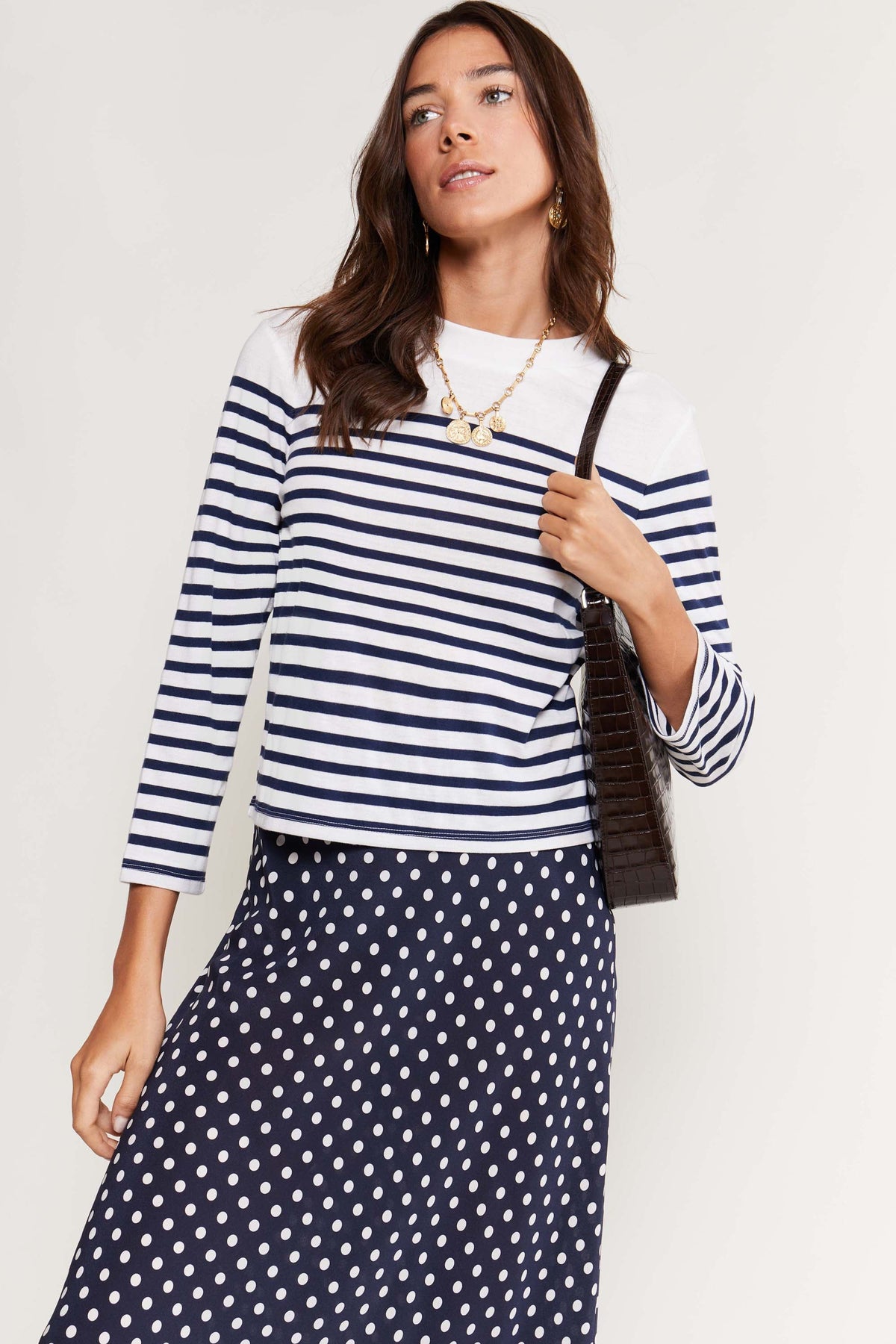 A woman wears the Francesco - Nautical Stripe Cream top paired with a navy polka dot skirt, necklace, and dark shoulder bag, standing against a plain background—showcasing effortlessly chic wardrobe staples.