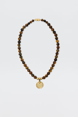 The Ischia - Brown necklace features round tiger's eye beads and a gold clasp, accented with a detailed circular gold pendant, shown on a plain white background.