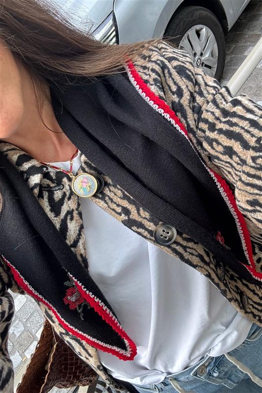 A person wears the Ari - Astro Sagittarius necklace over a white shirt with a leopard print cardigan, red trim, black scarf, and round colorful pin. A car and cobblestone ground appear in the background.