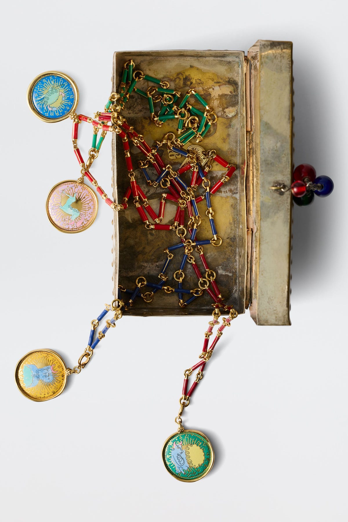 Ari - Astro Sagittarius: A partially open tarnished metal jewelry box reveals a zodiac necklace with vibrant gold enamel Sagittarius pendant, statement disc charms, and red, green, and blue geometric chain links spilling onto a white surface.