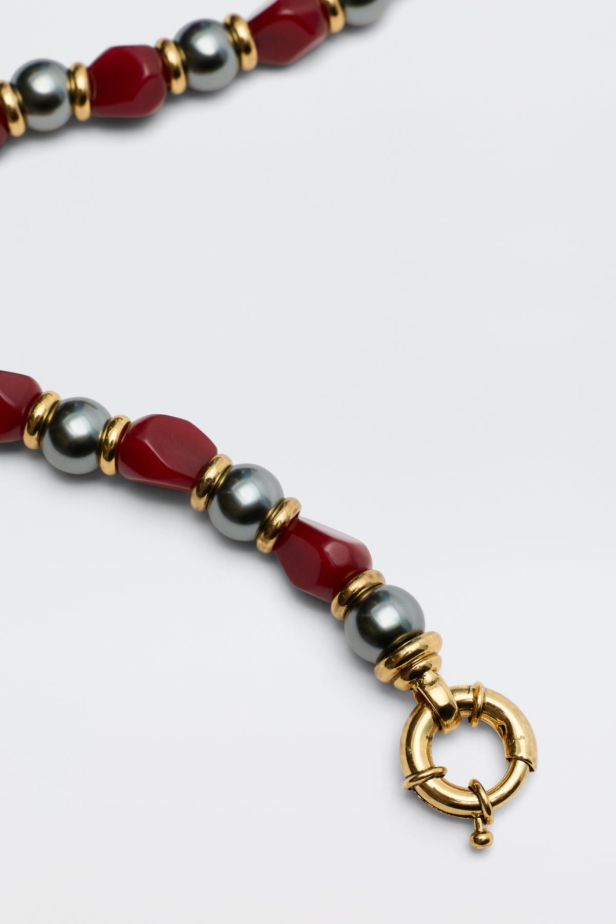 A close-up of the Palmara - Burgundy necklace, showcasing alternating red and metallic grey beads with gold accents and a round gold clasp on a plain white background.