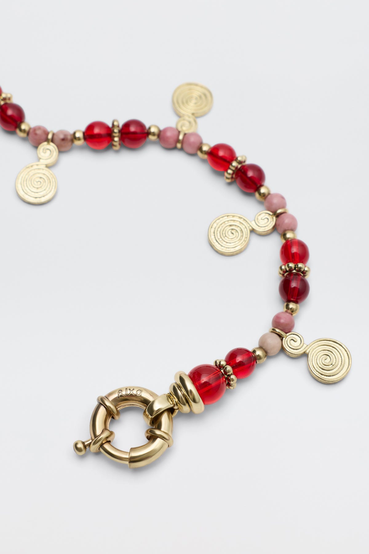 Tala - Burgundy: A beaded necklace with burgundy and pink beads, gold spacer beads, gold spiral charms, and a large round gold clasp, displayed on a light background.